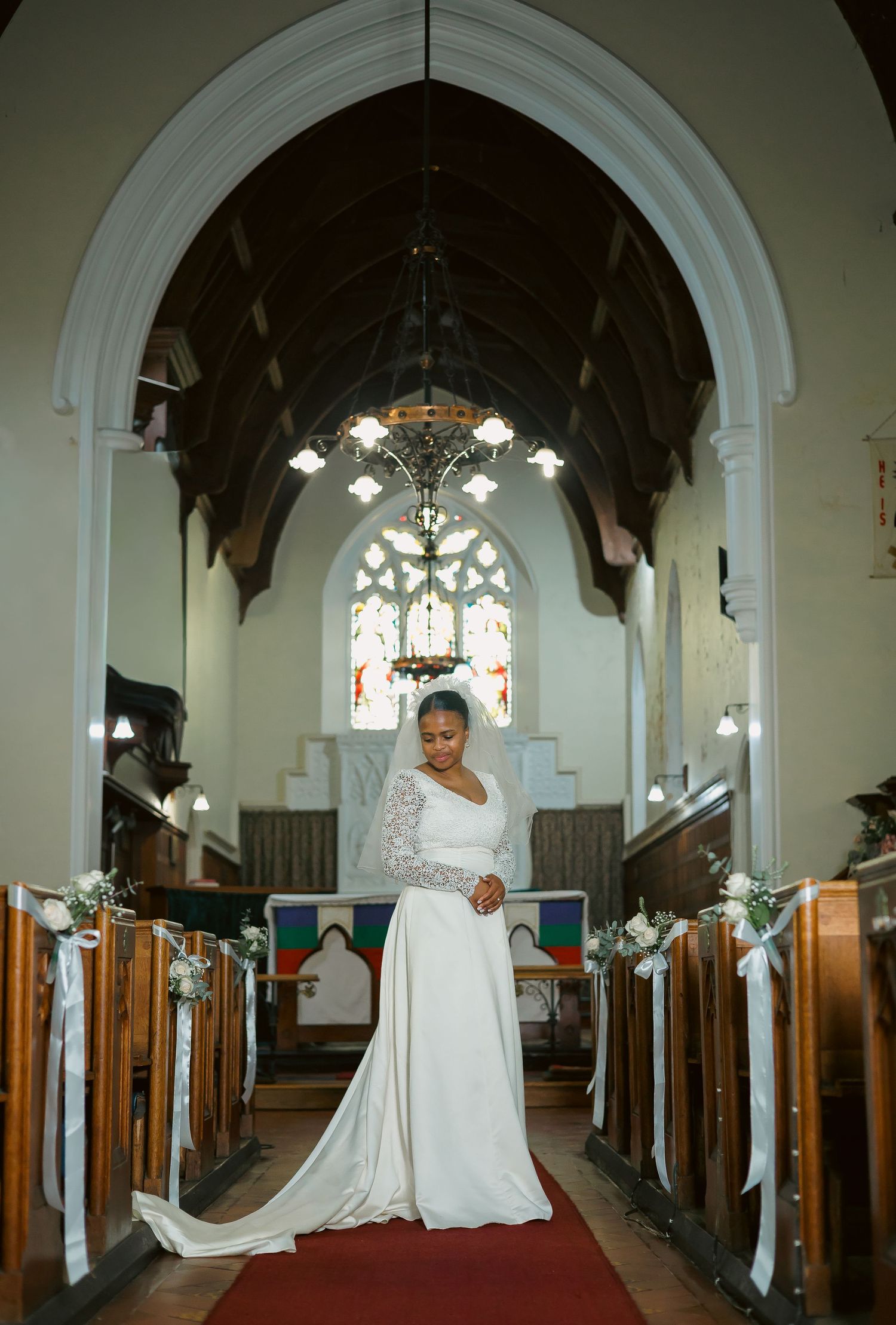 Elegant bride in long white gown with veil stands in church aisle.