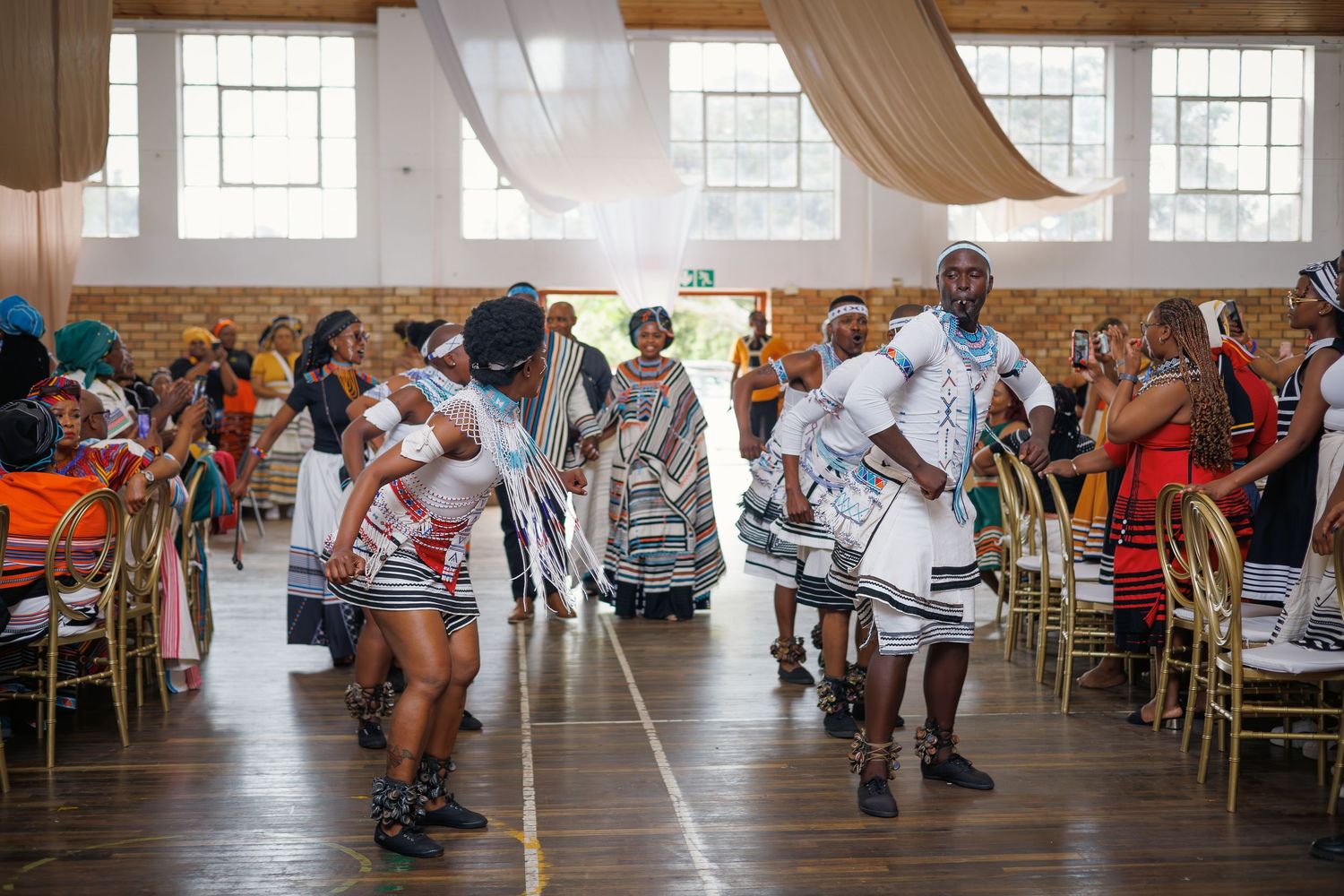 A group of performers in traditional Xhosa attire dance energetically inside a decorated hall at a celebration.