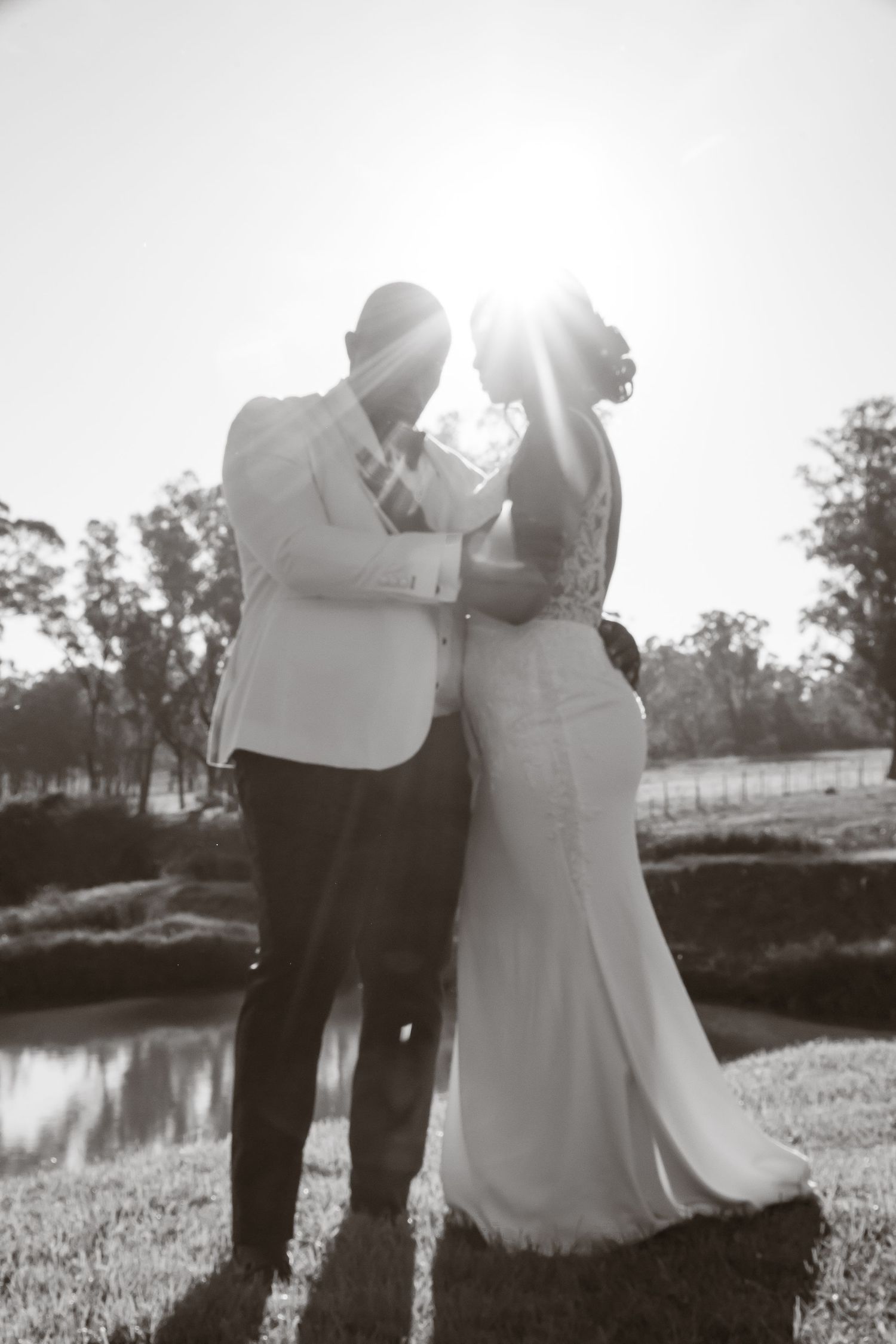 Newlyweds share a romantic moment backlit by golden sunlight near a serene pond on their wedding day.