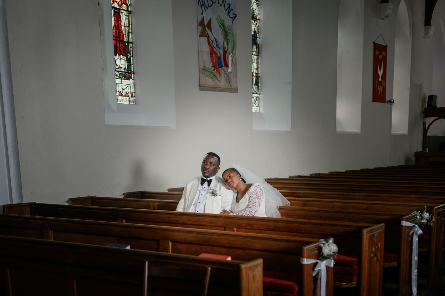 Bride and groom sitting closely together in a sunlit church pew surrounded by stained glass windows.