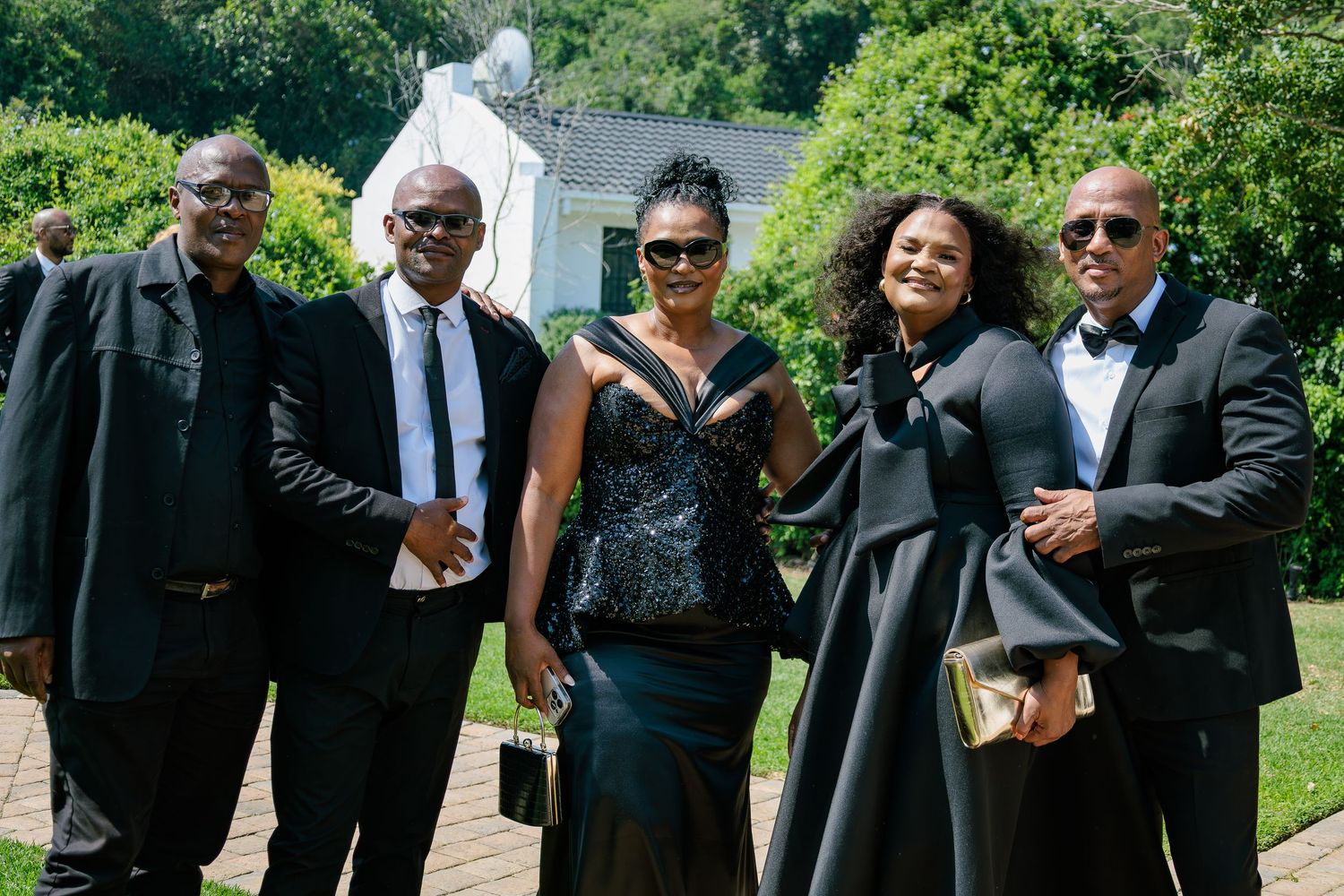 Group of five elegantly dressed wedding guests posing together outdoors in black formal attire.