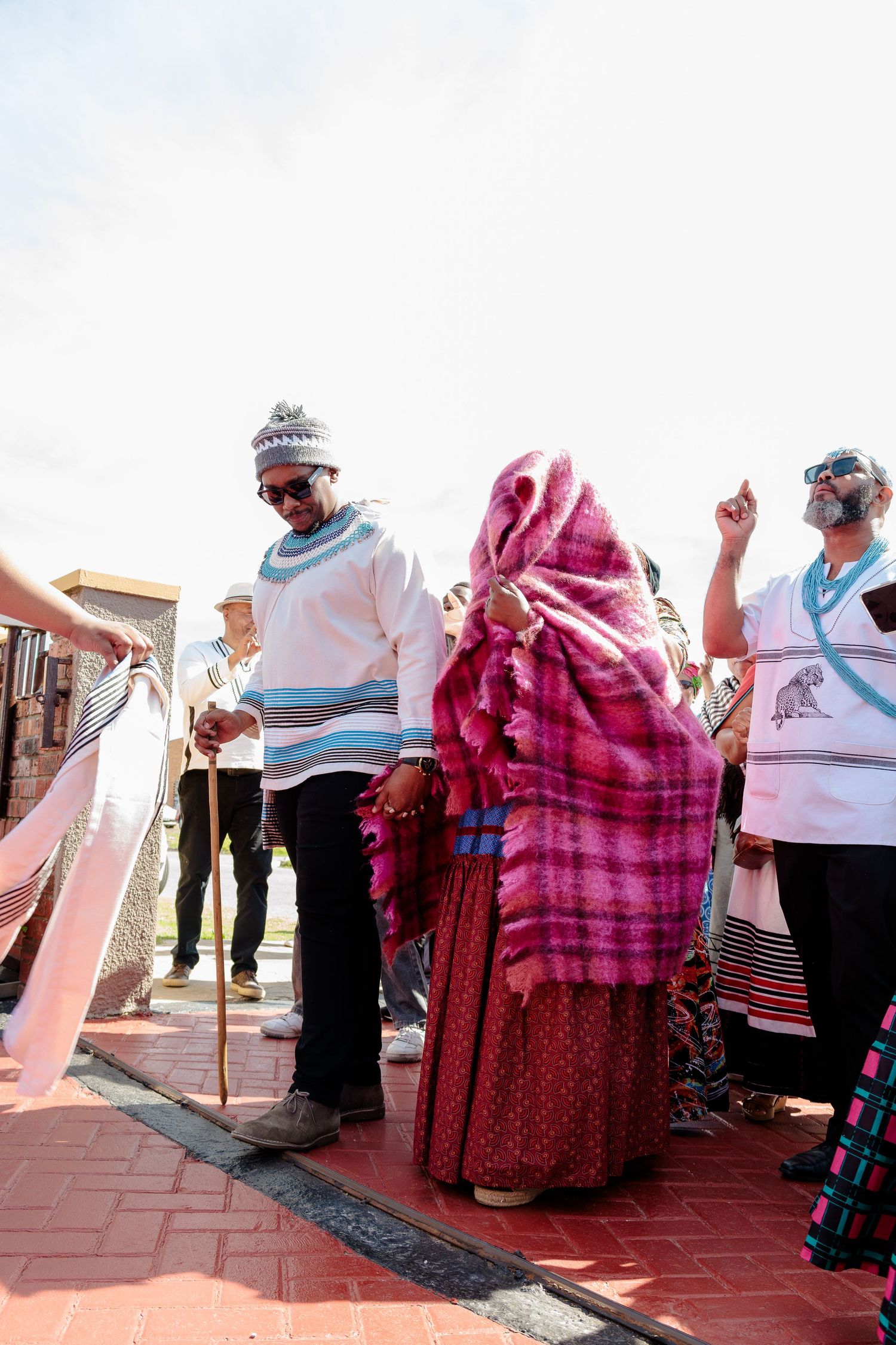 Man in white Xhosa blanket assists woman in pink blanket during traditional ceremony outdoors.