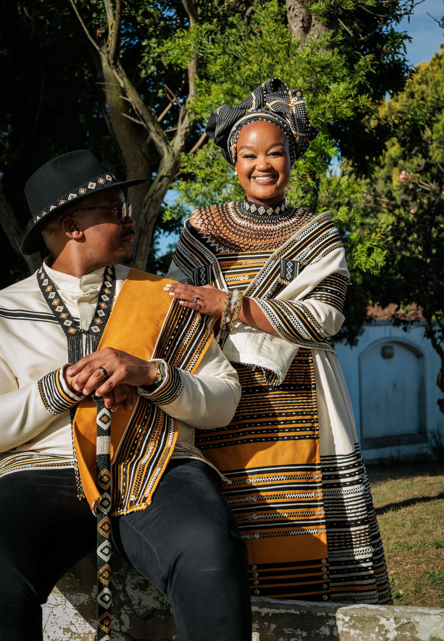 Smiling couple in traditional Xhosa attire seated outdoors, woman in beaded headpiece laughing joyfully.