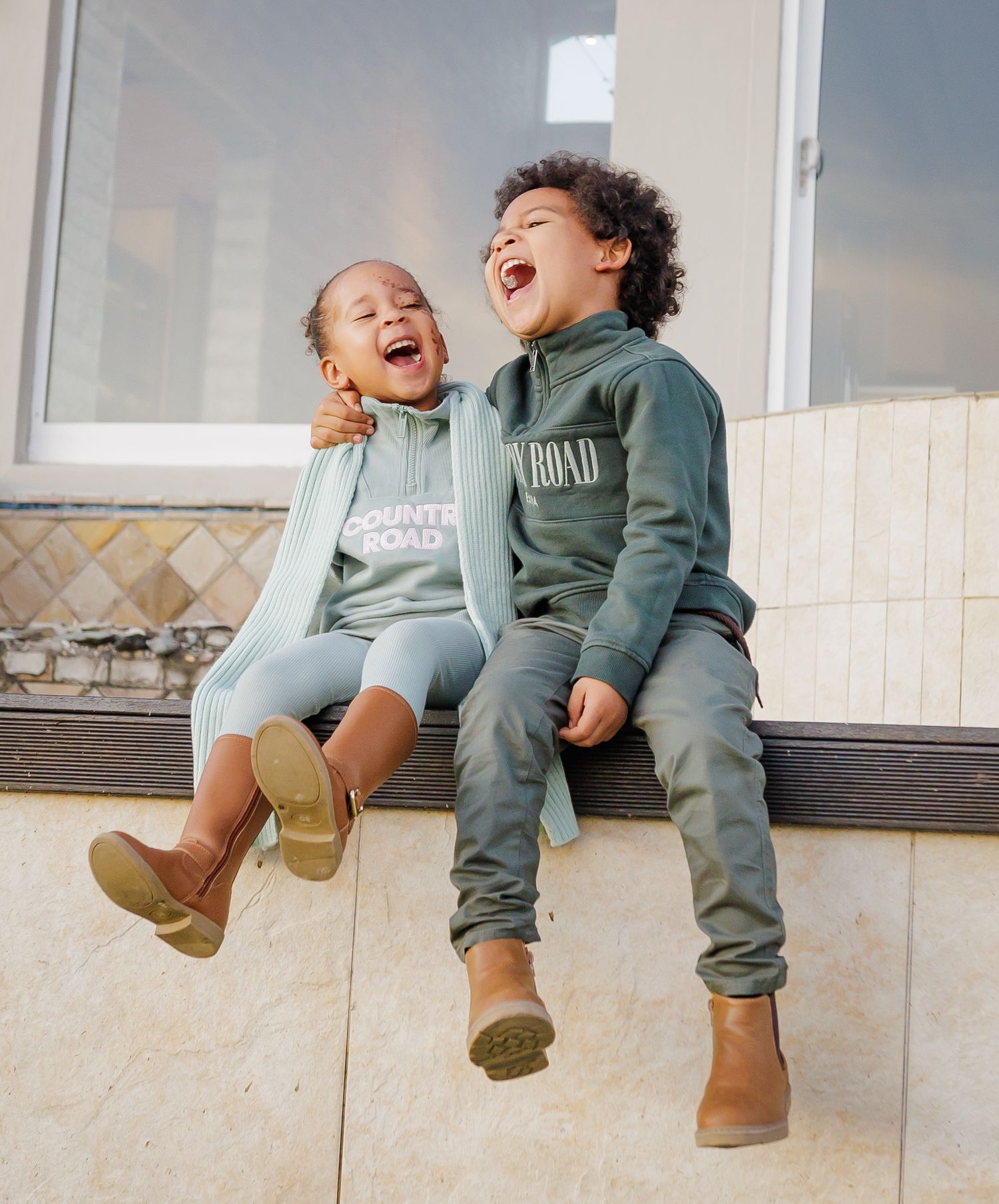 Two young children laughing joyfully while sitting together wearing matching Country Road hoodies.