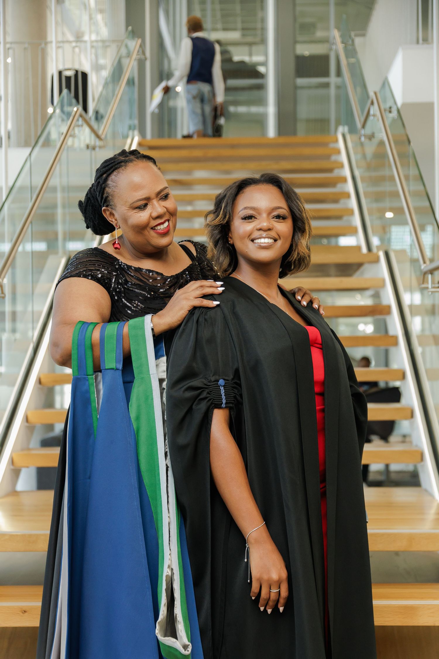 A Black mother and her daughter in graduation gown smile together on a staircase inside a modern venue.