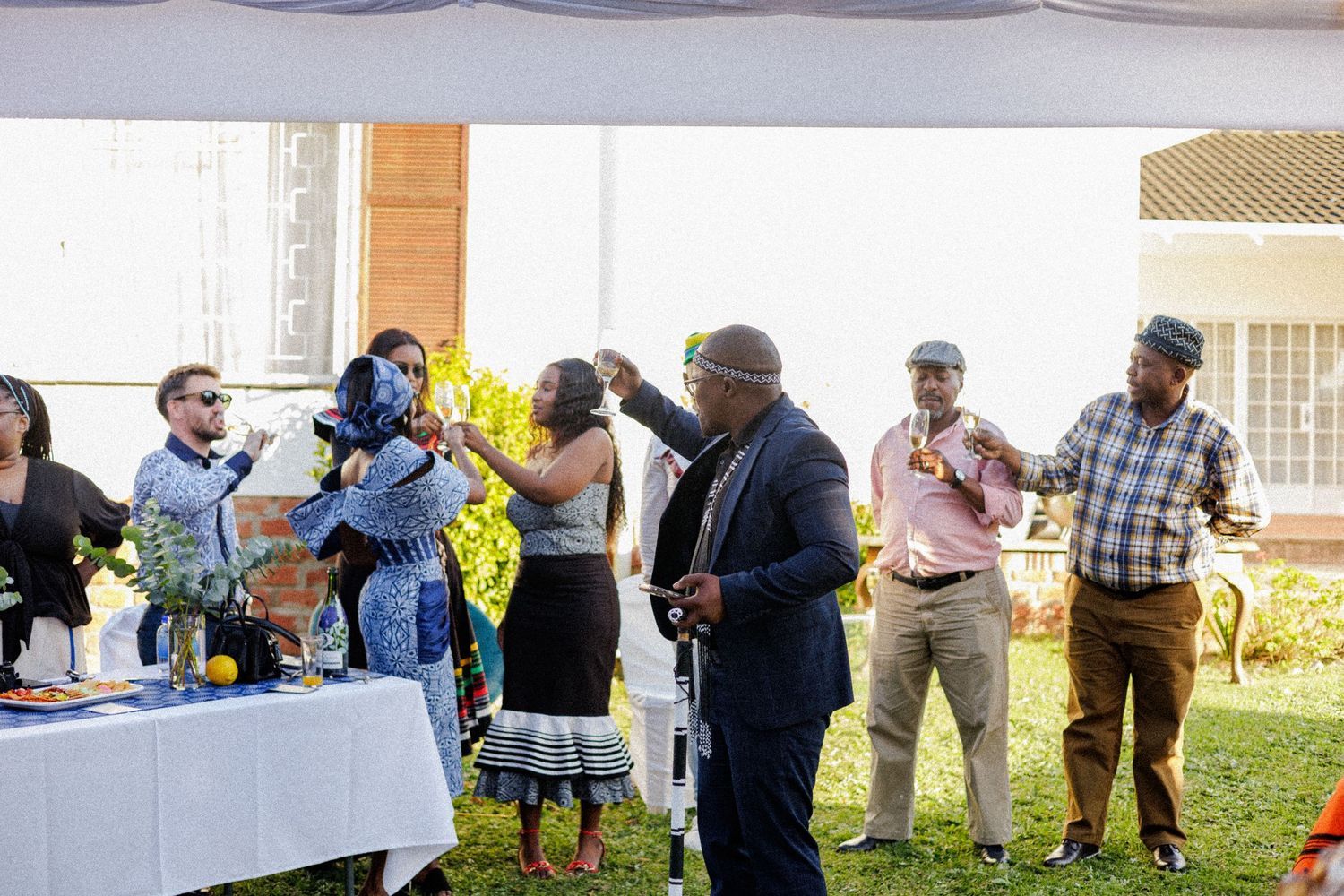 Group of guests gathered under tent at outdoor African cultural celebration with drinks and conversation.