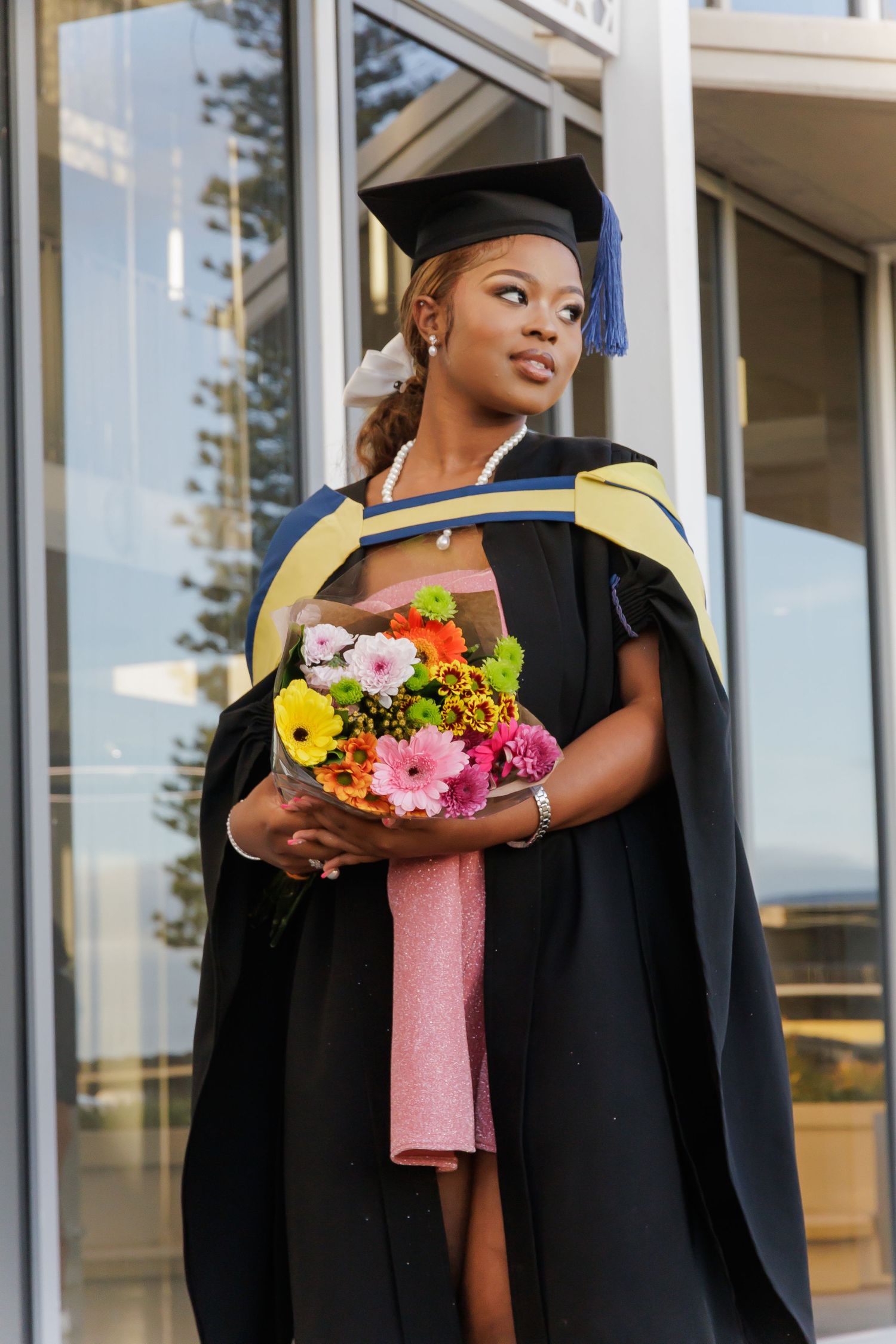 A Black female graduate in black cap and gown holds a colorful bouquet of flowers outside a building.