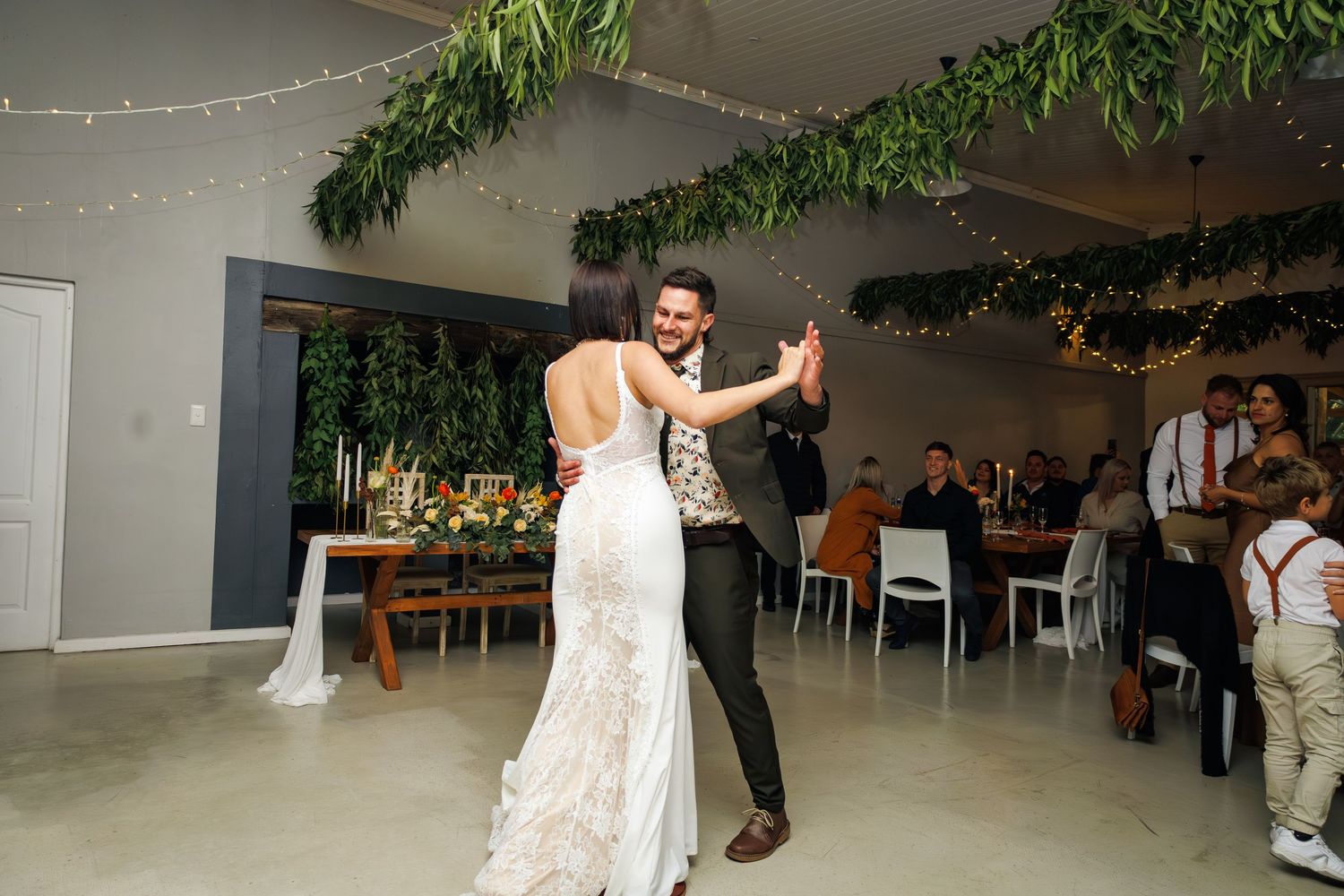 Bride and groom share their first dance in decorated wedding venue with greenery and string lights.