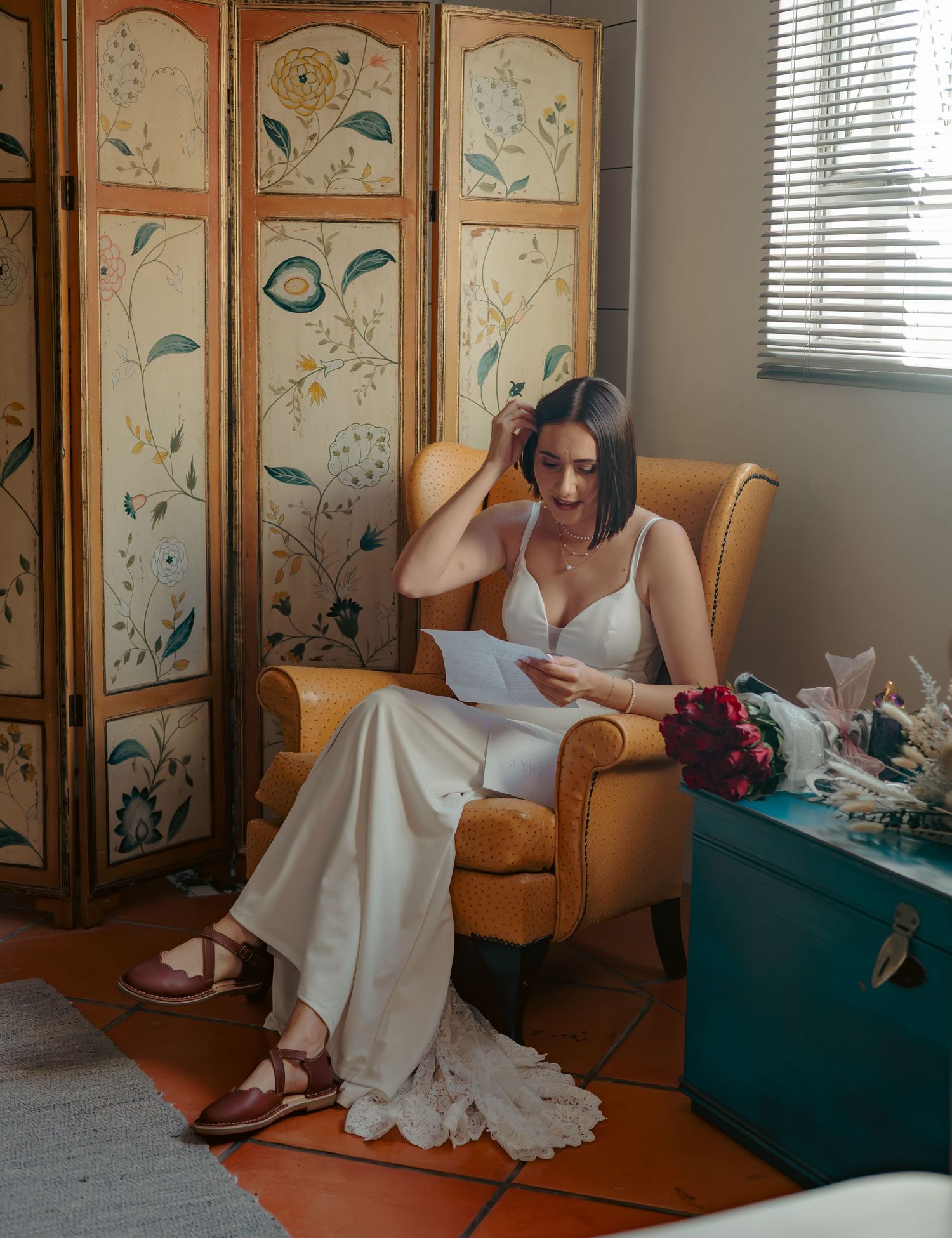 Bride in white gown reading letter in yellow armchair beside floral decorative screen.