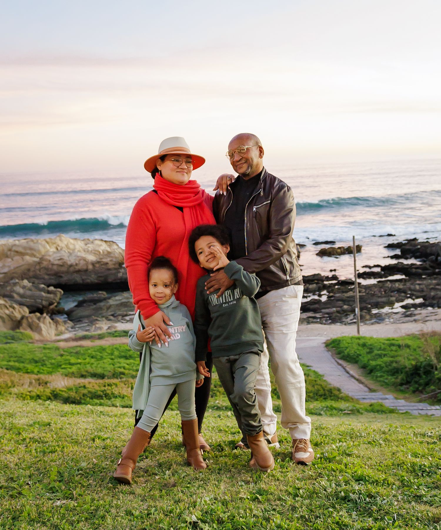 Smiling family of four posing together on a scenic coastal lawn during a beautiful golden sunset.