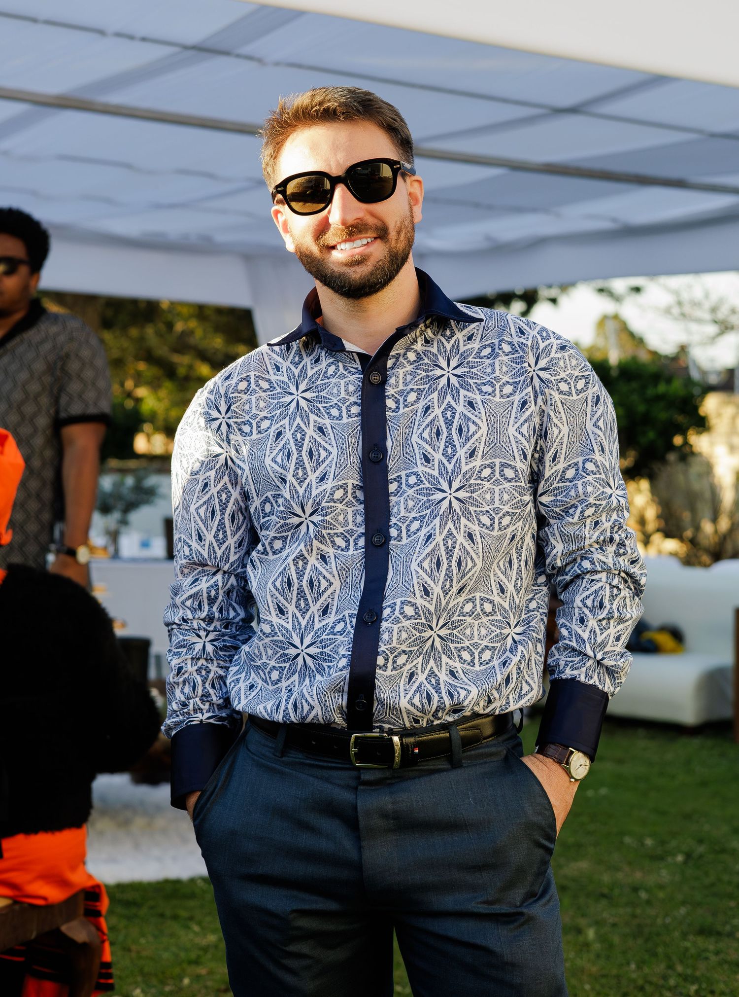 Smiling man in blue patterned shirt and sunglasses posing at an outdoor cultural celebration event.