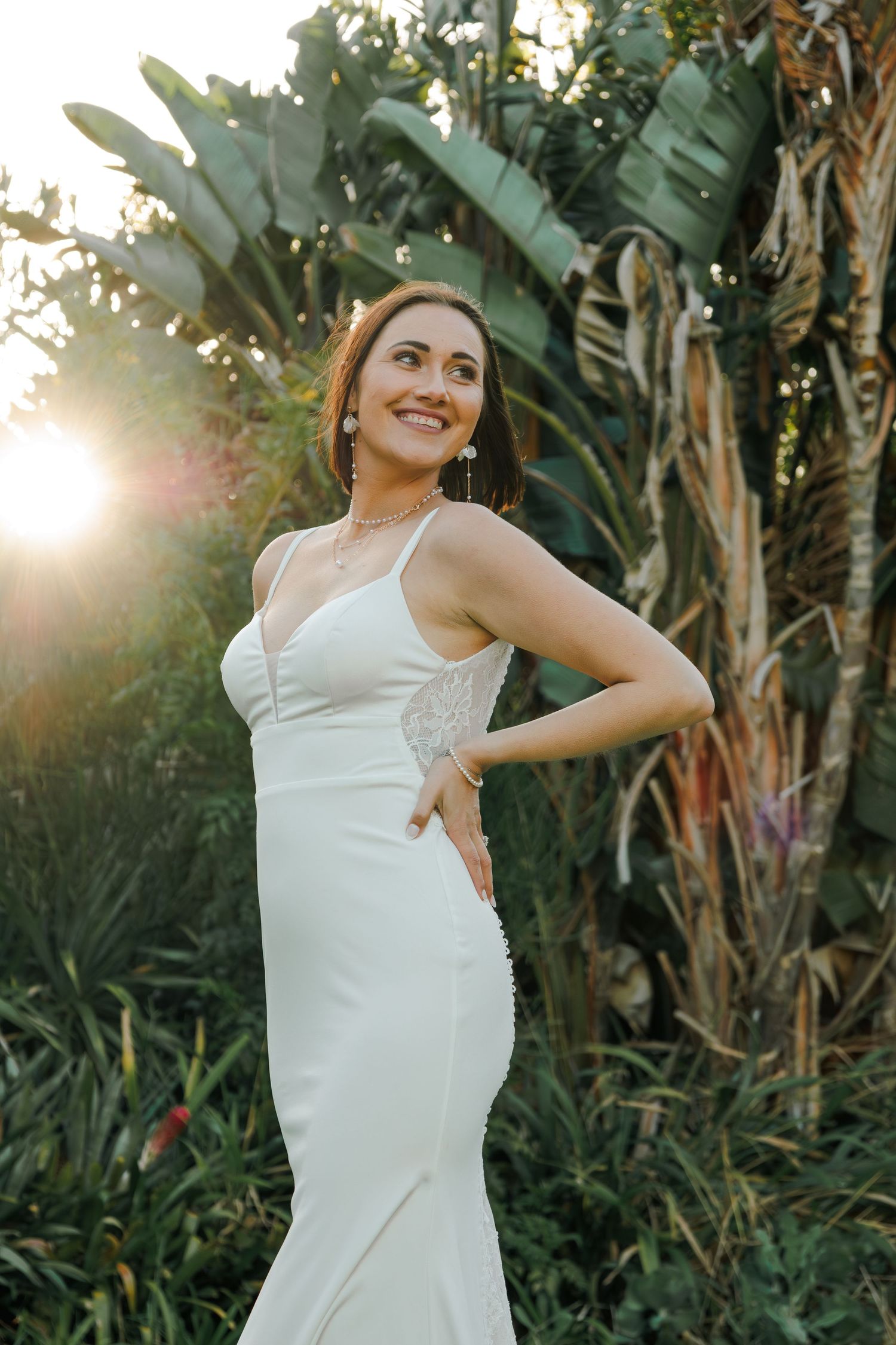 Radiant bride in fitted white gown posing confidently in tropical garden sunset.