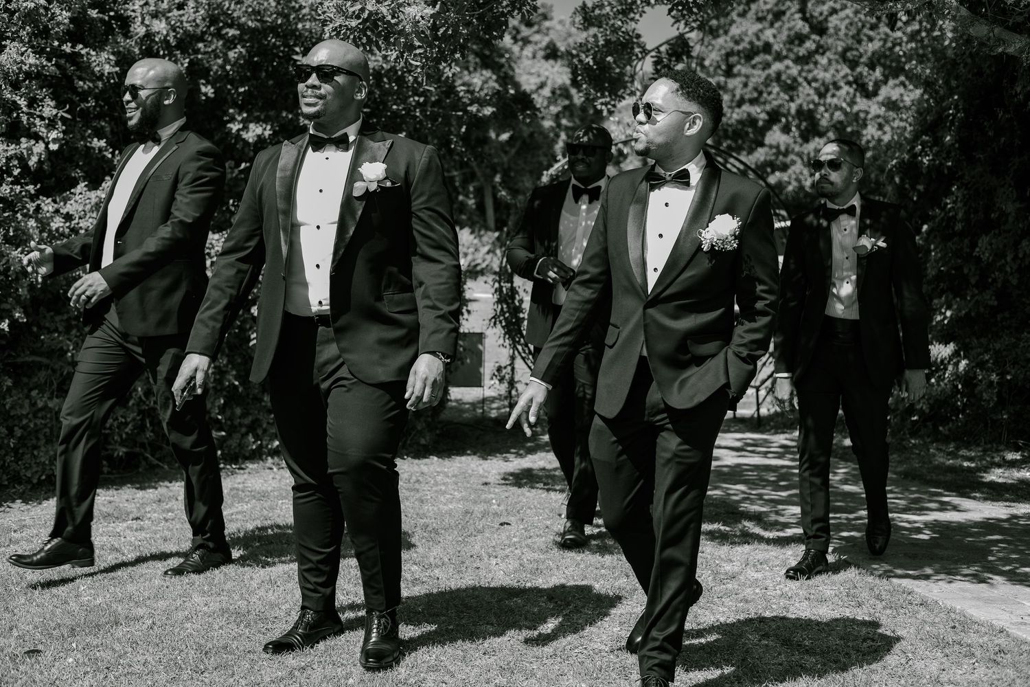 Groomsmen in black tuxedos walking confidently across a sunlit garden in a black and white wedding photo.