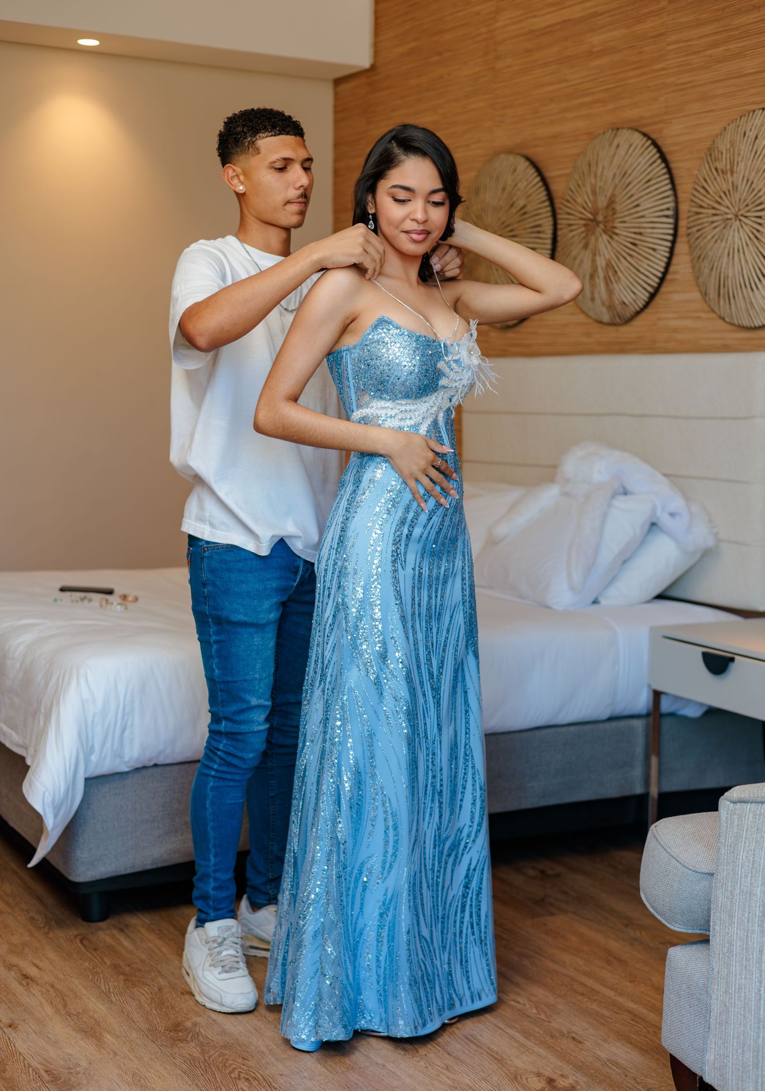 Young man helping woman in a sparkling blue sequin strapless prom gown in a modern hotel room.