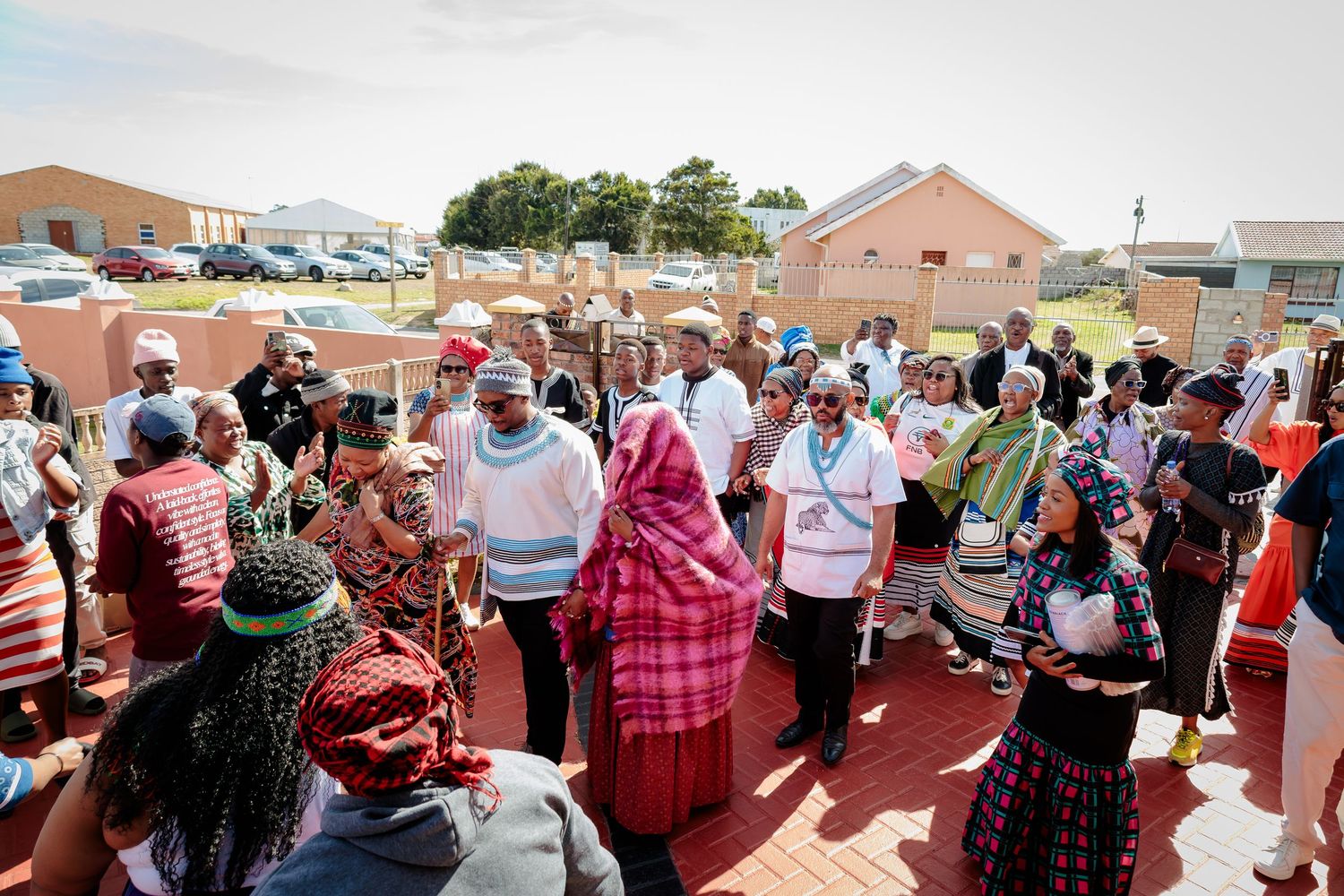 A large group in traditional African clothing gathers outdoors in a residential neighborhood during a ceremony.