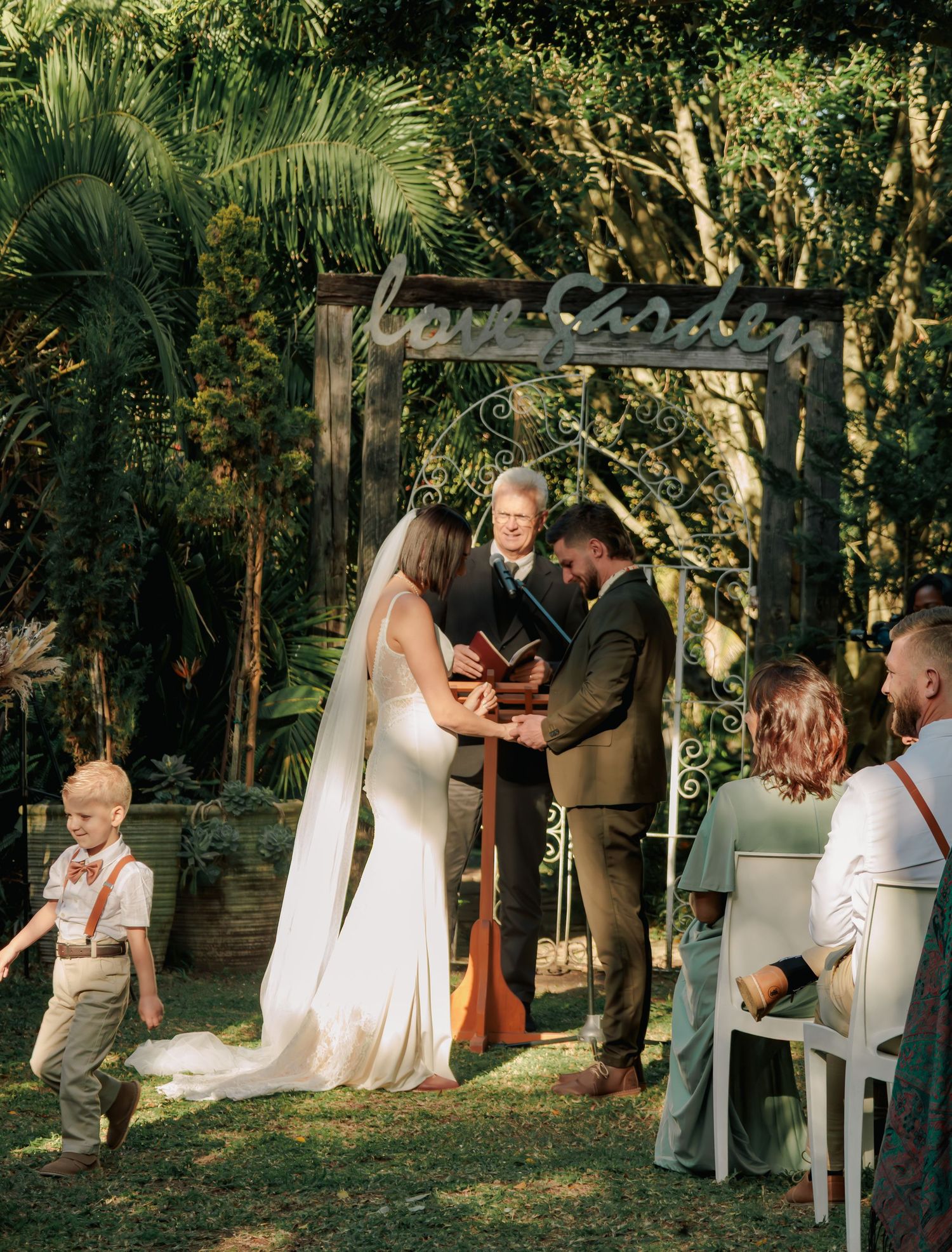 Couple holding hands during outdoor garden wedding ceremony under a wooden arch with guests seated in white chairs.