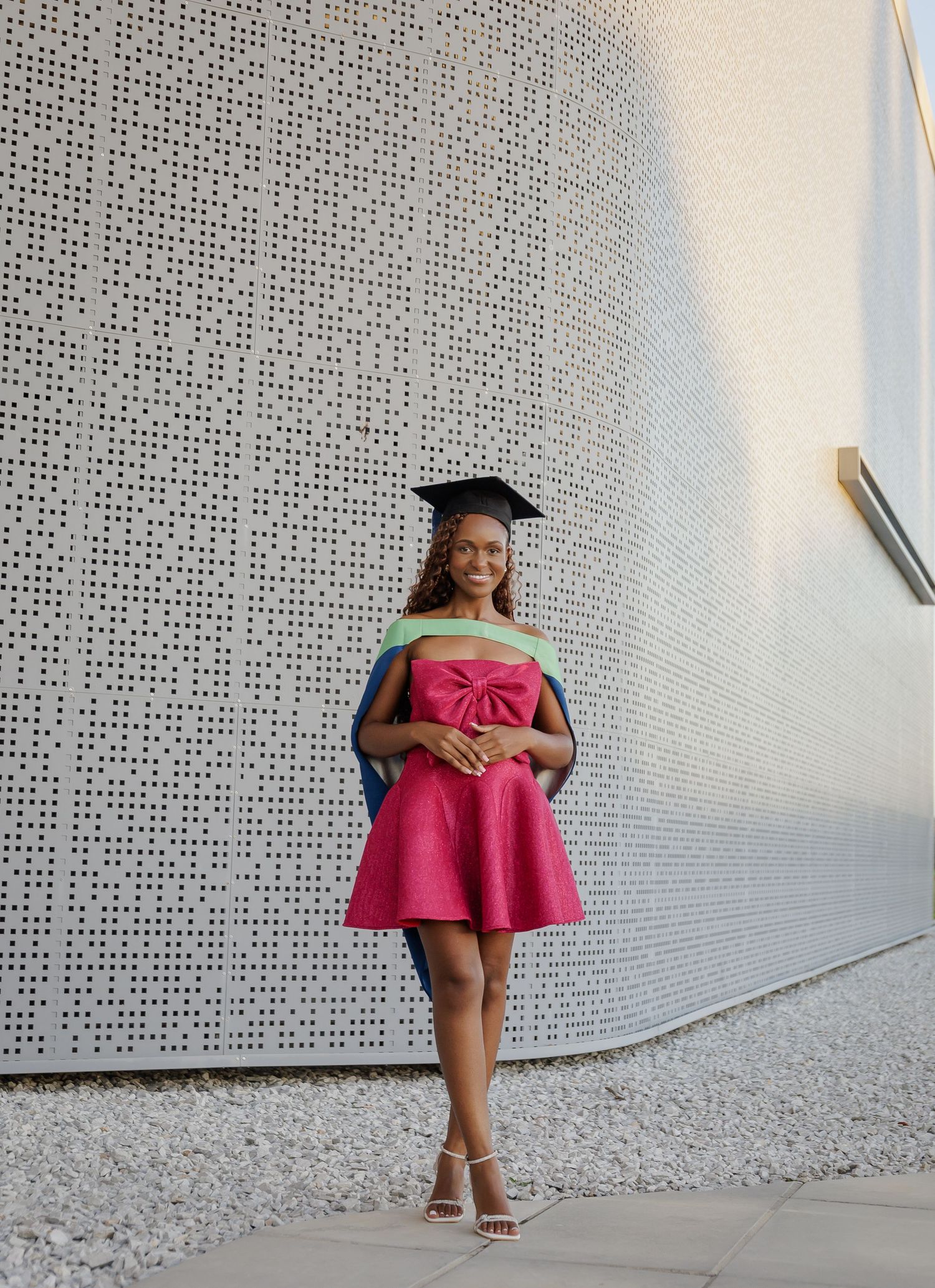A young Black woman in a pink dress and graduation cap poses confidently against a textured white wall.
