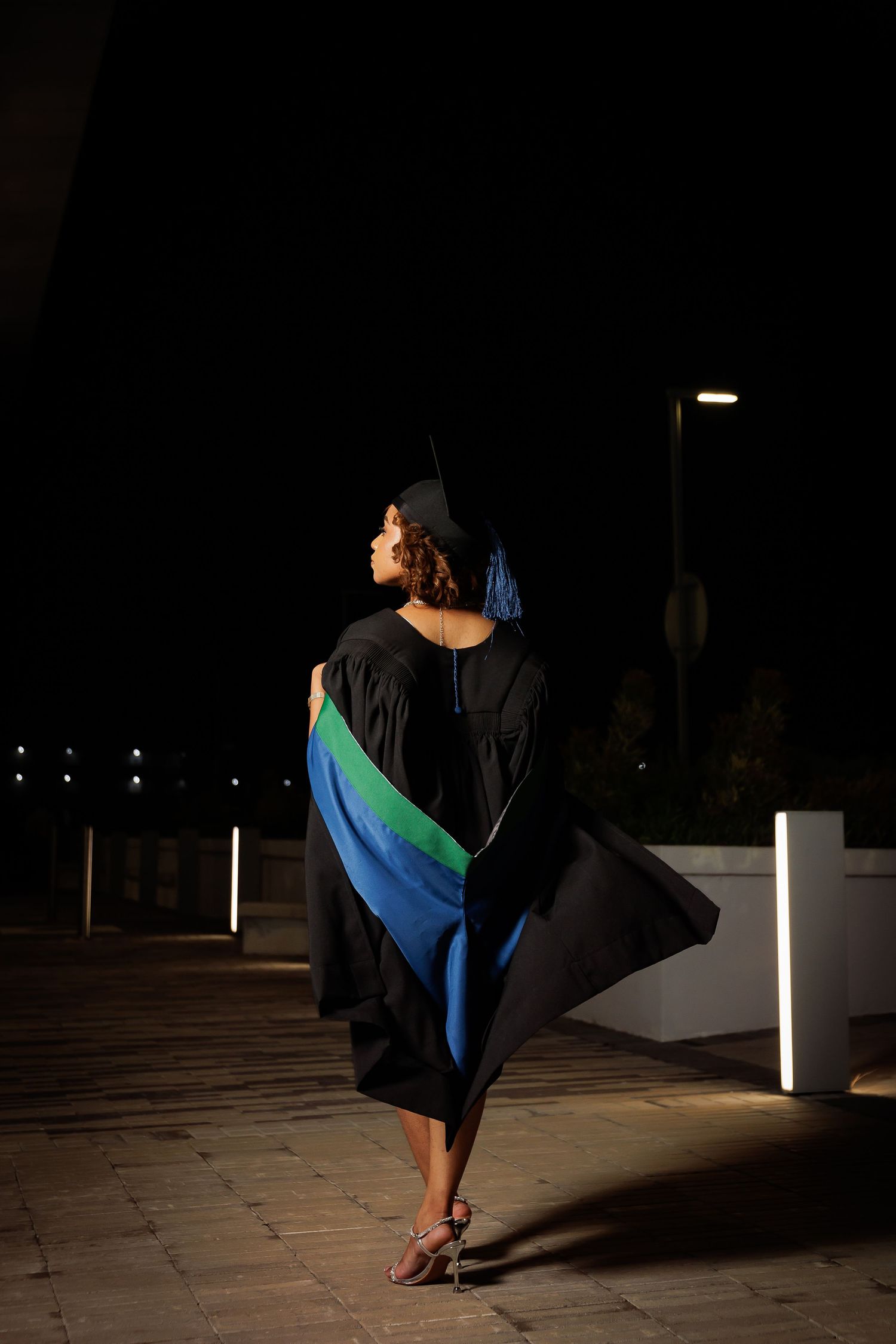 Graduate in black gown and cap with blue and green hood walks outdoors at night.