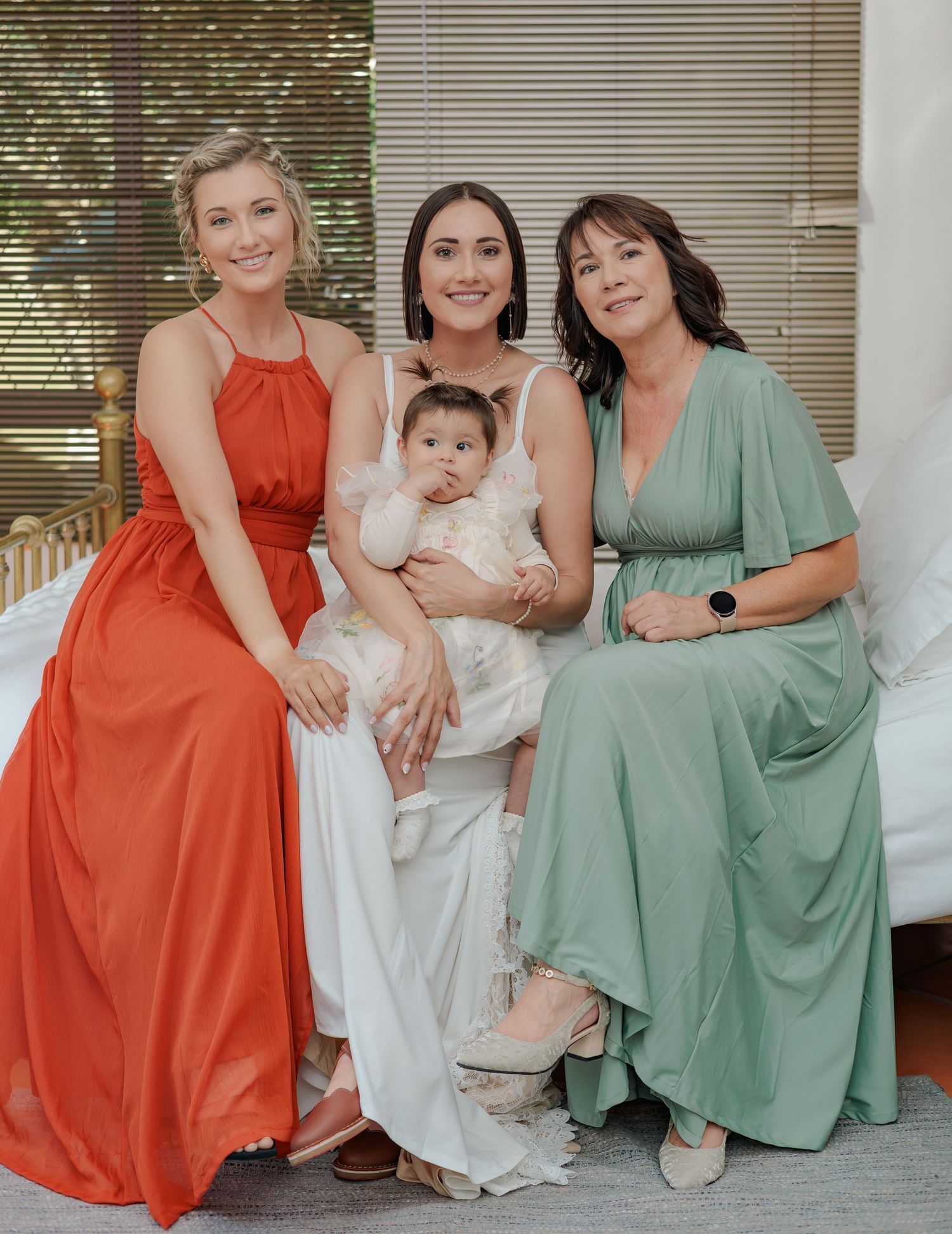 Bride in white dress holding baby seated with bridesmaid in orange and woman in sage green dress.