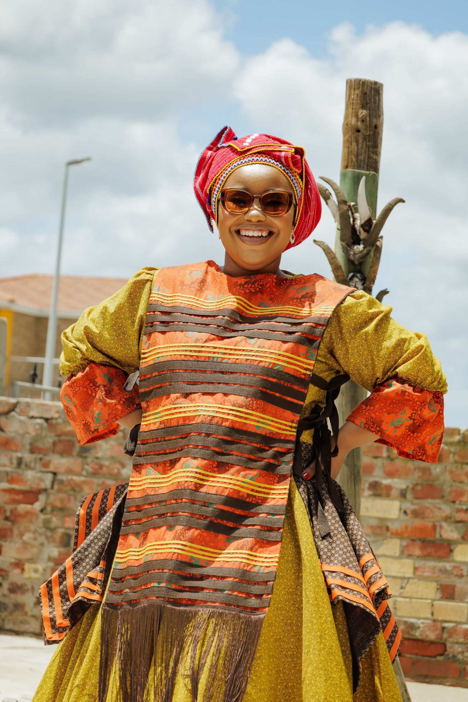 Joyful woman in vibrant traditional African clothing and sunglasses posing confidently outdoors on a sunny day.
