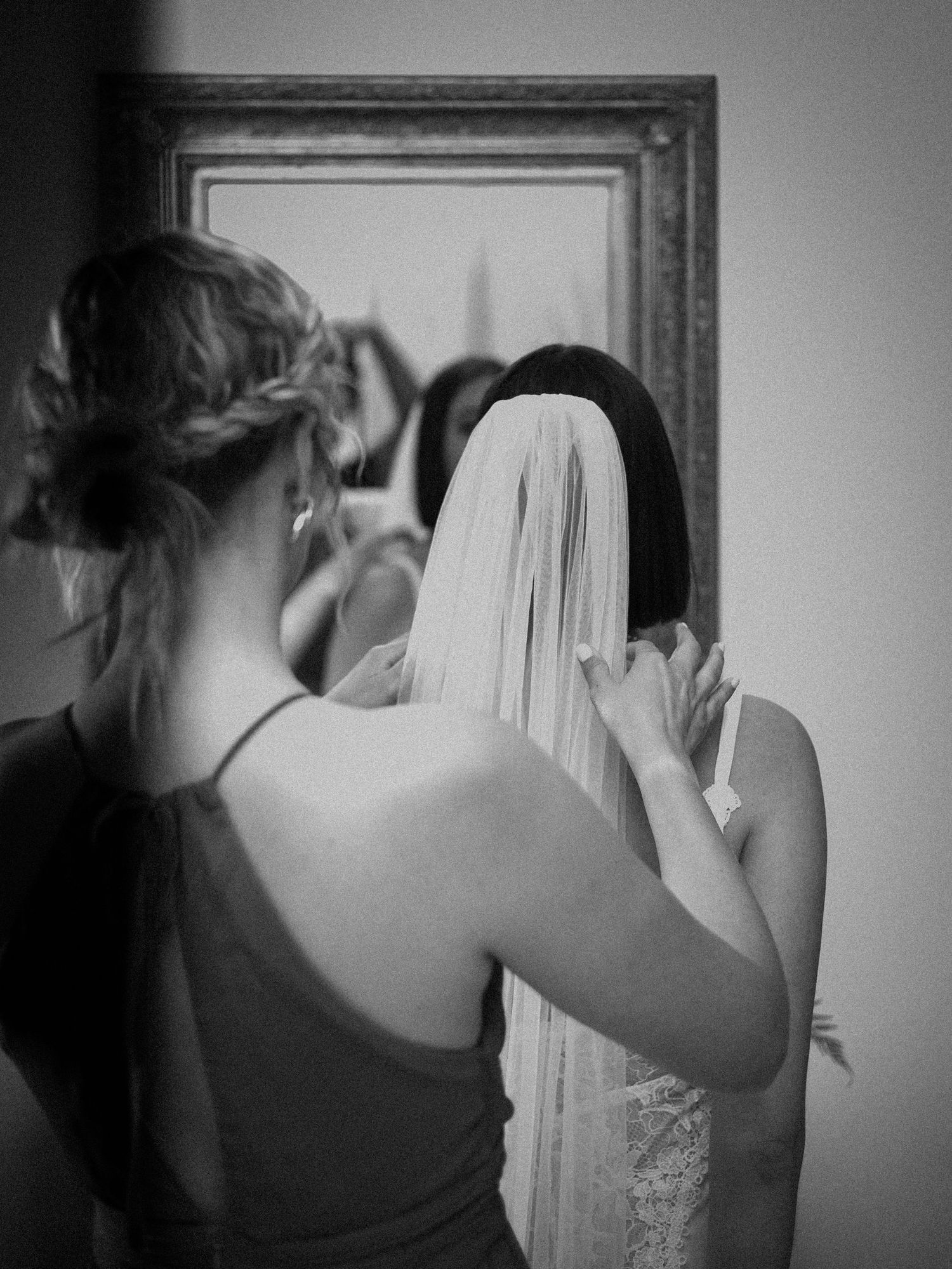Black and white photo of bridesmaid with braided updo placing veil on bride before mirror.