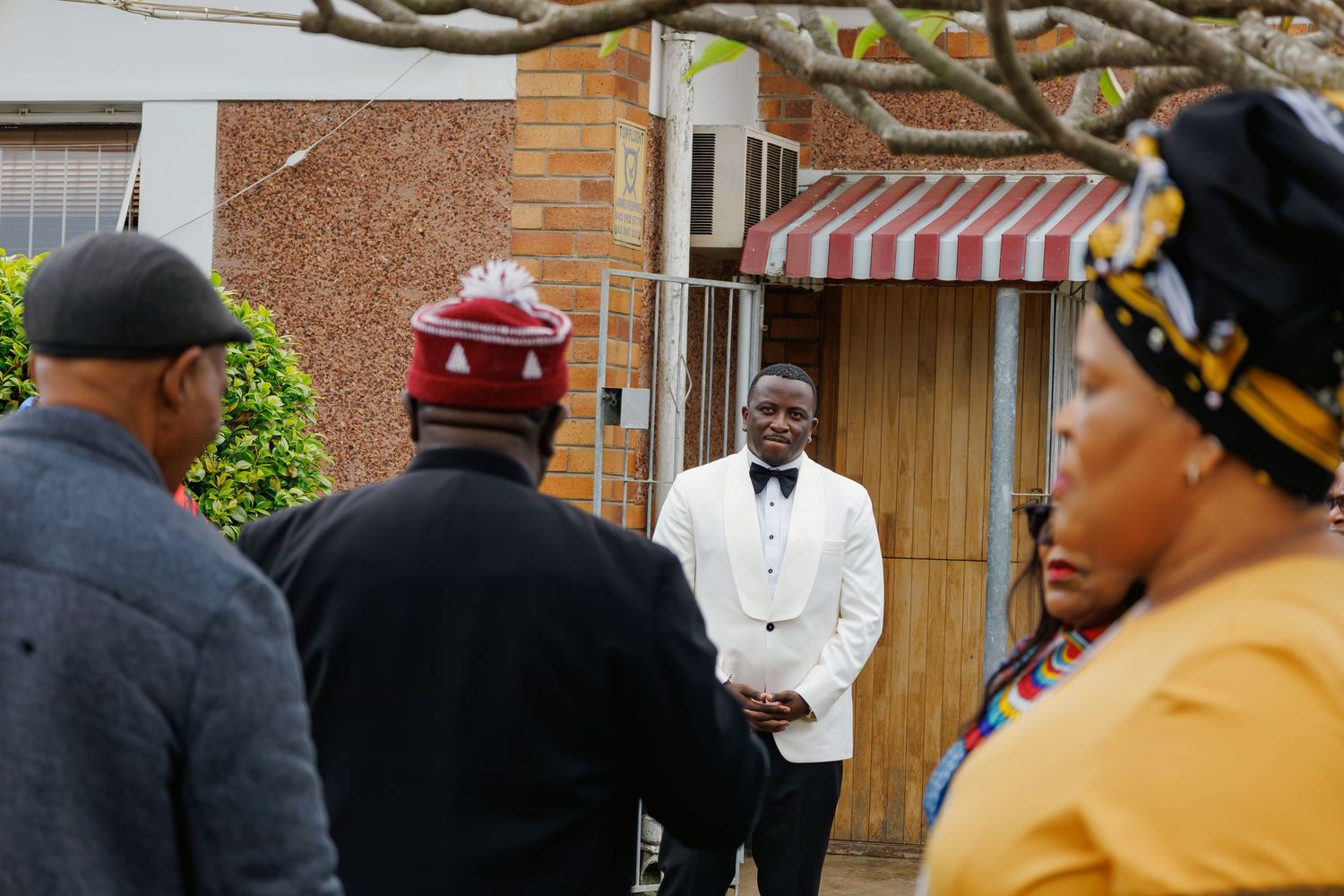 Man in white suit greets guests in traditional African attire outside a residential home.