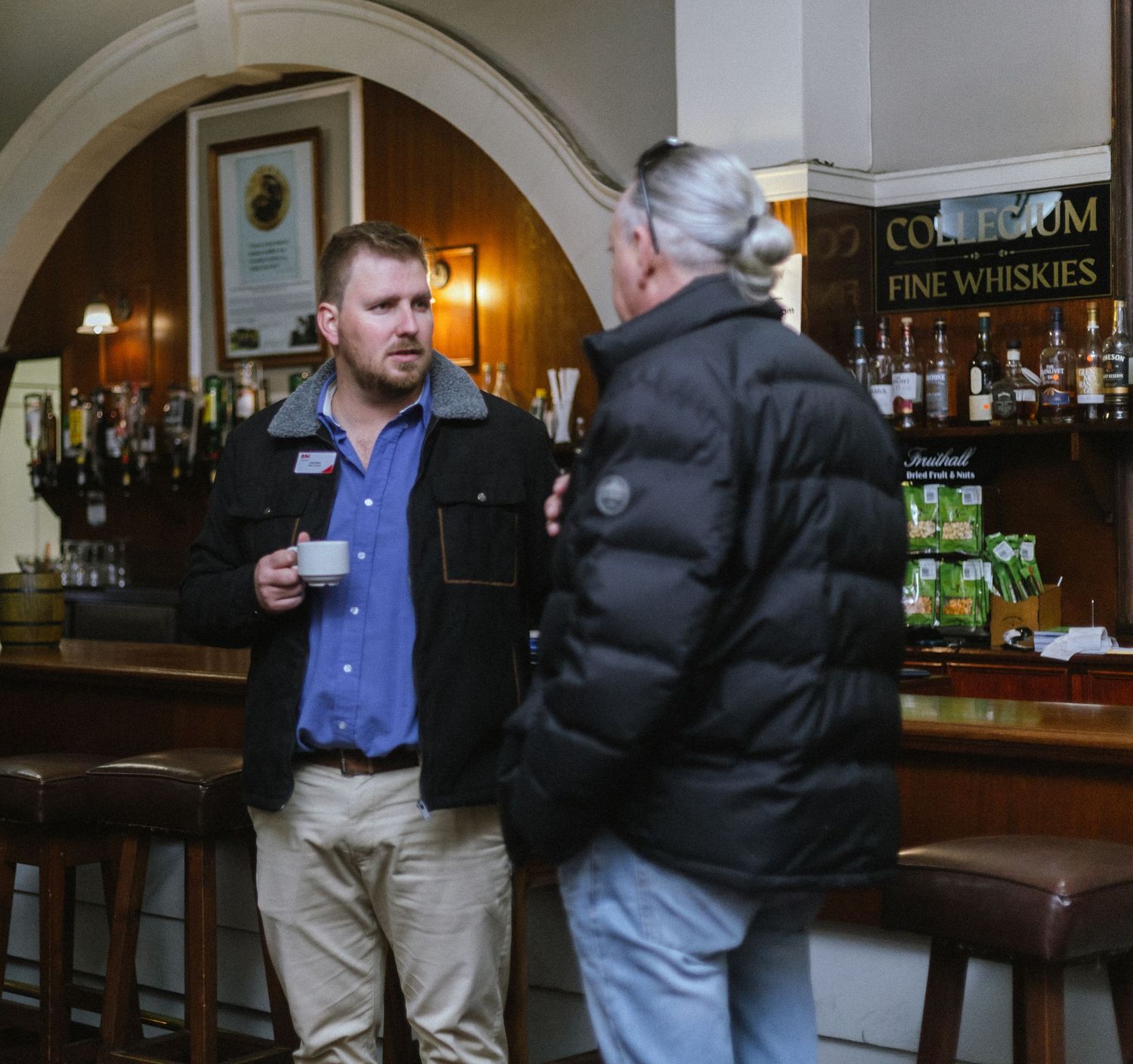 Two men in casual attire having a conversation near a bar with fine whisky bottles in background.
