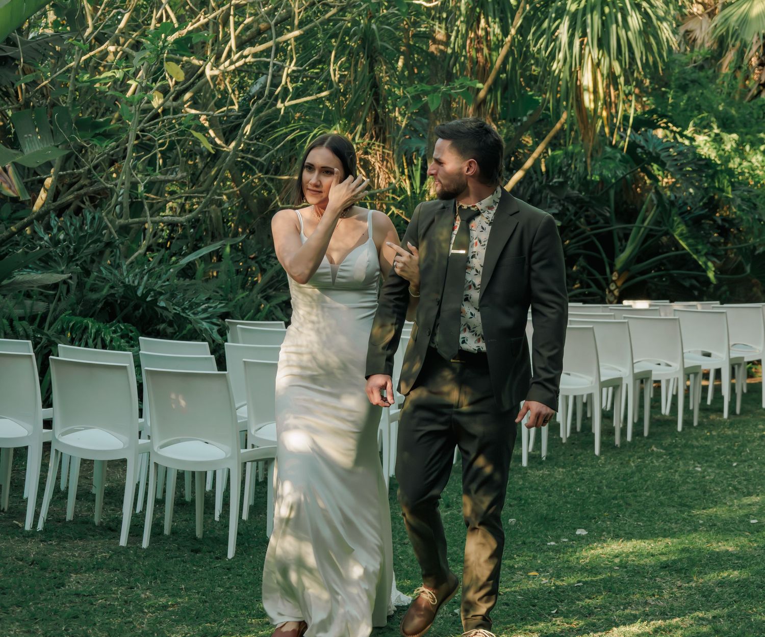 Emotional bride wiping tears while walking with groom through sunlit garden after outdoor wedding ceremony.
