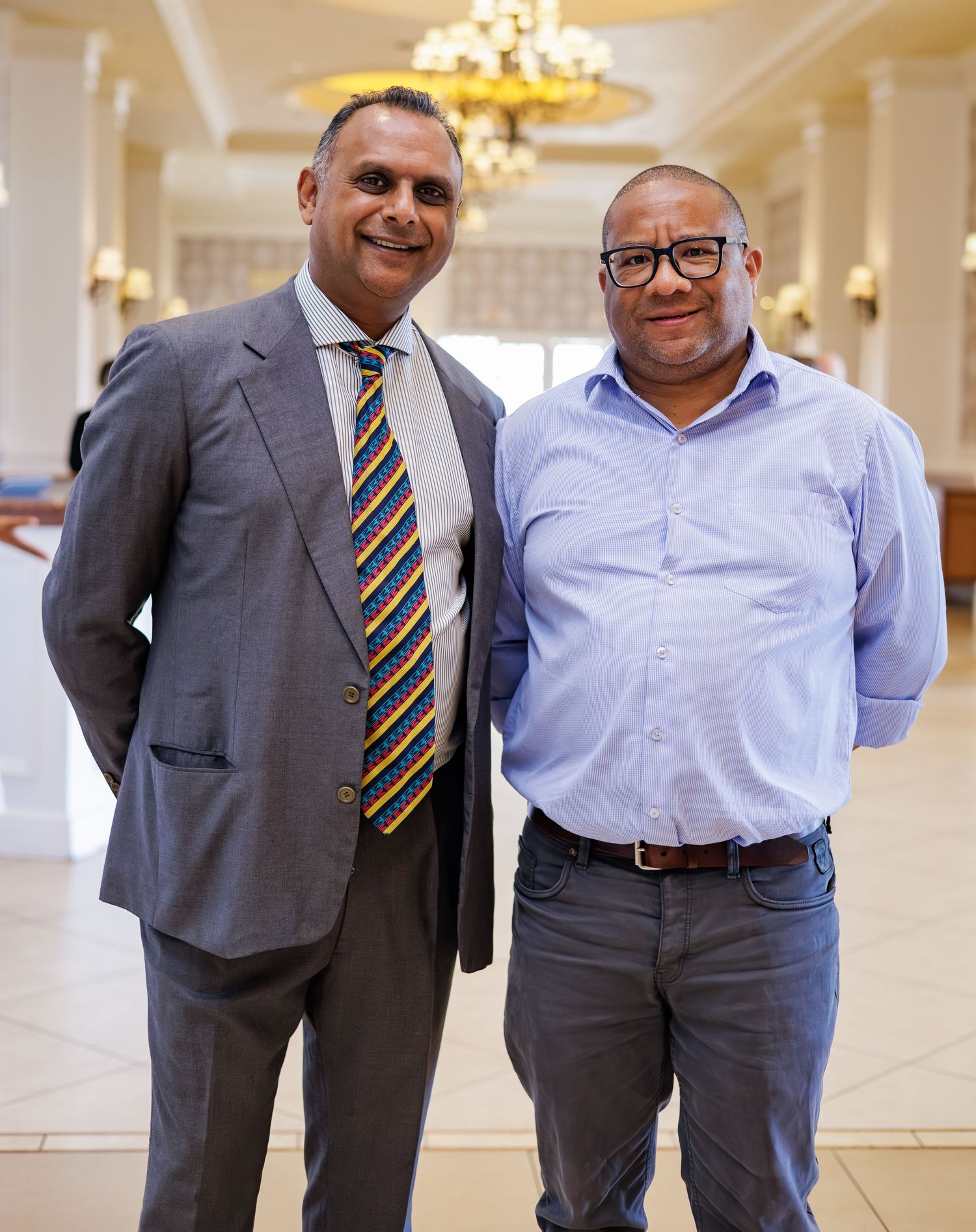 Two professionally dressed men smiling together in an elegant chandelier-lit banquet hall.