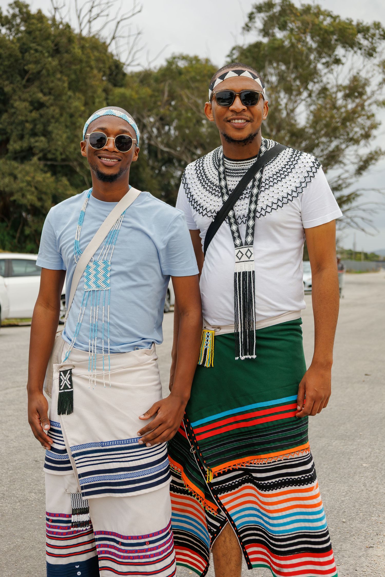 Two young men smile wearing traditional Xhosa attire with striped wraps and beaded accessories outdoors.
