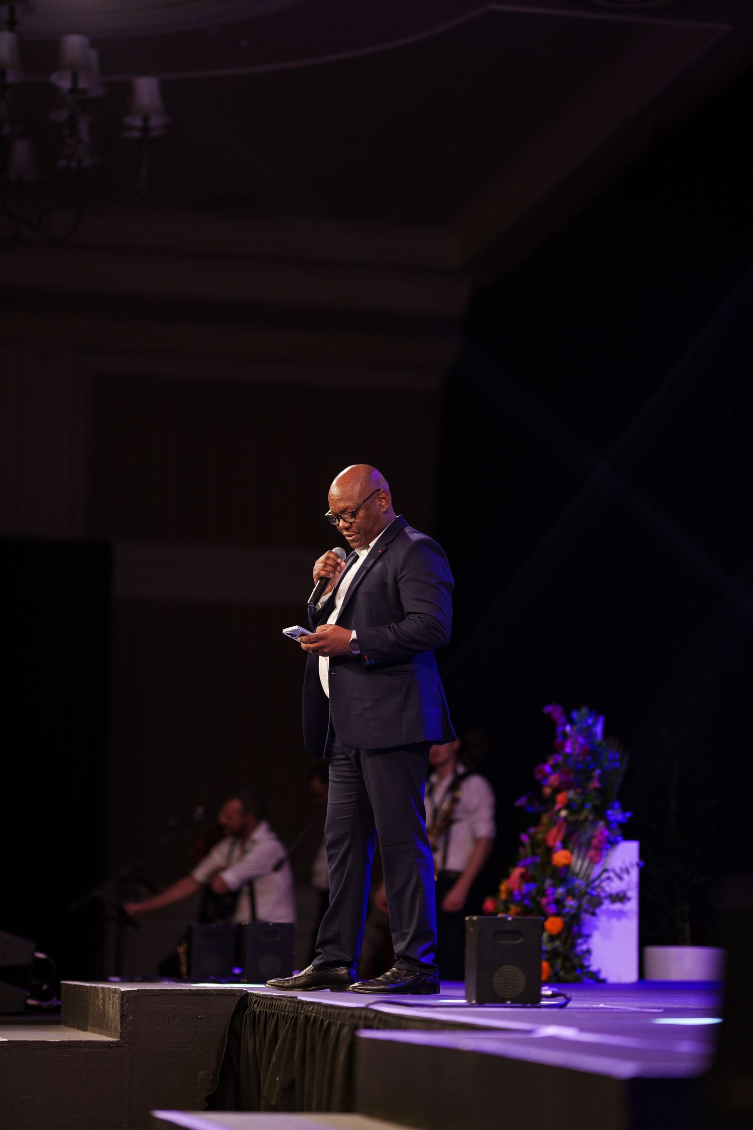 Man in grey suit speaking on stage with microphone at elegant indoor event venue.