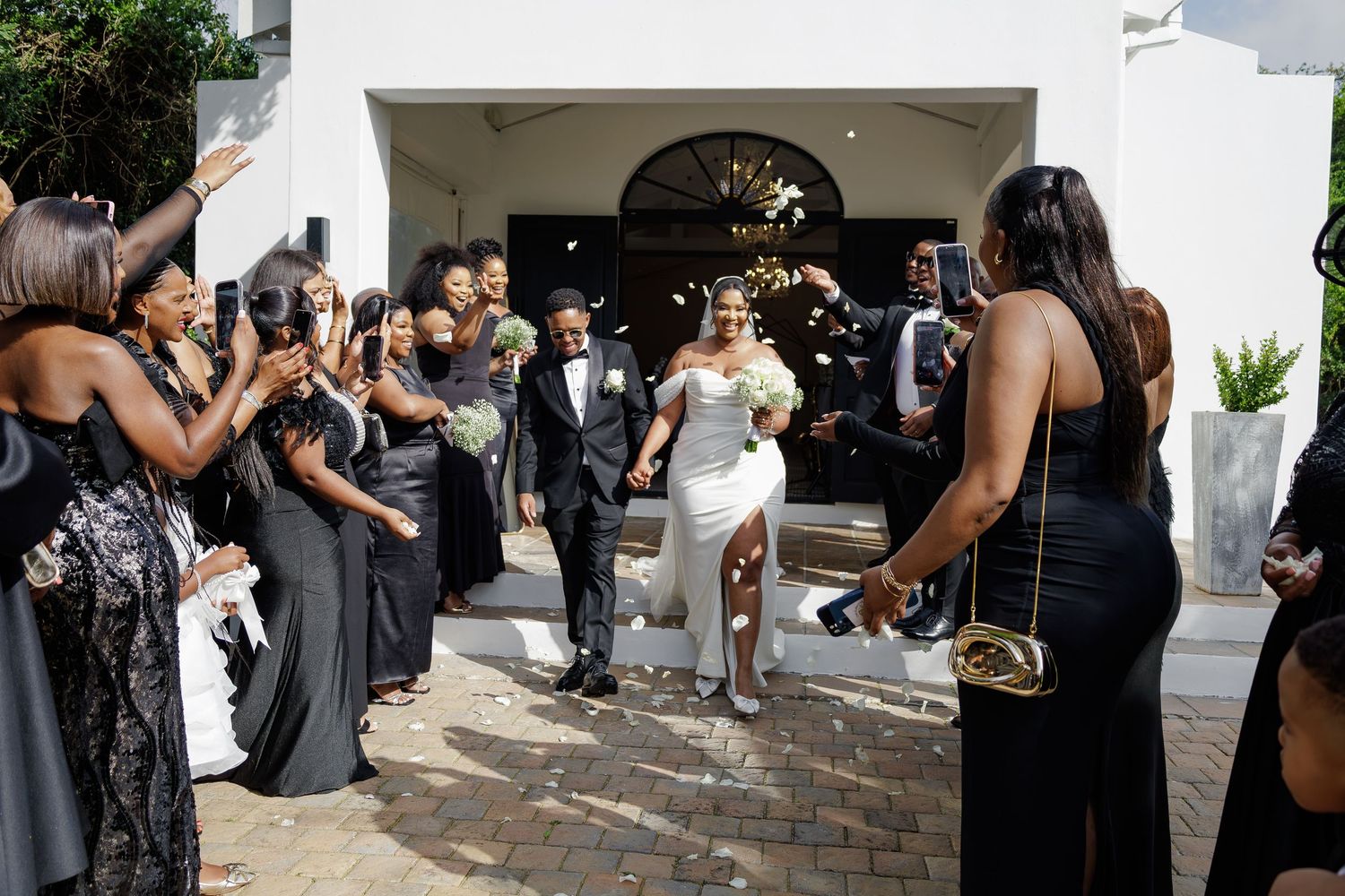 Newlywed couple walks through applauding bridal party outside elegant white venue.