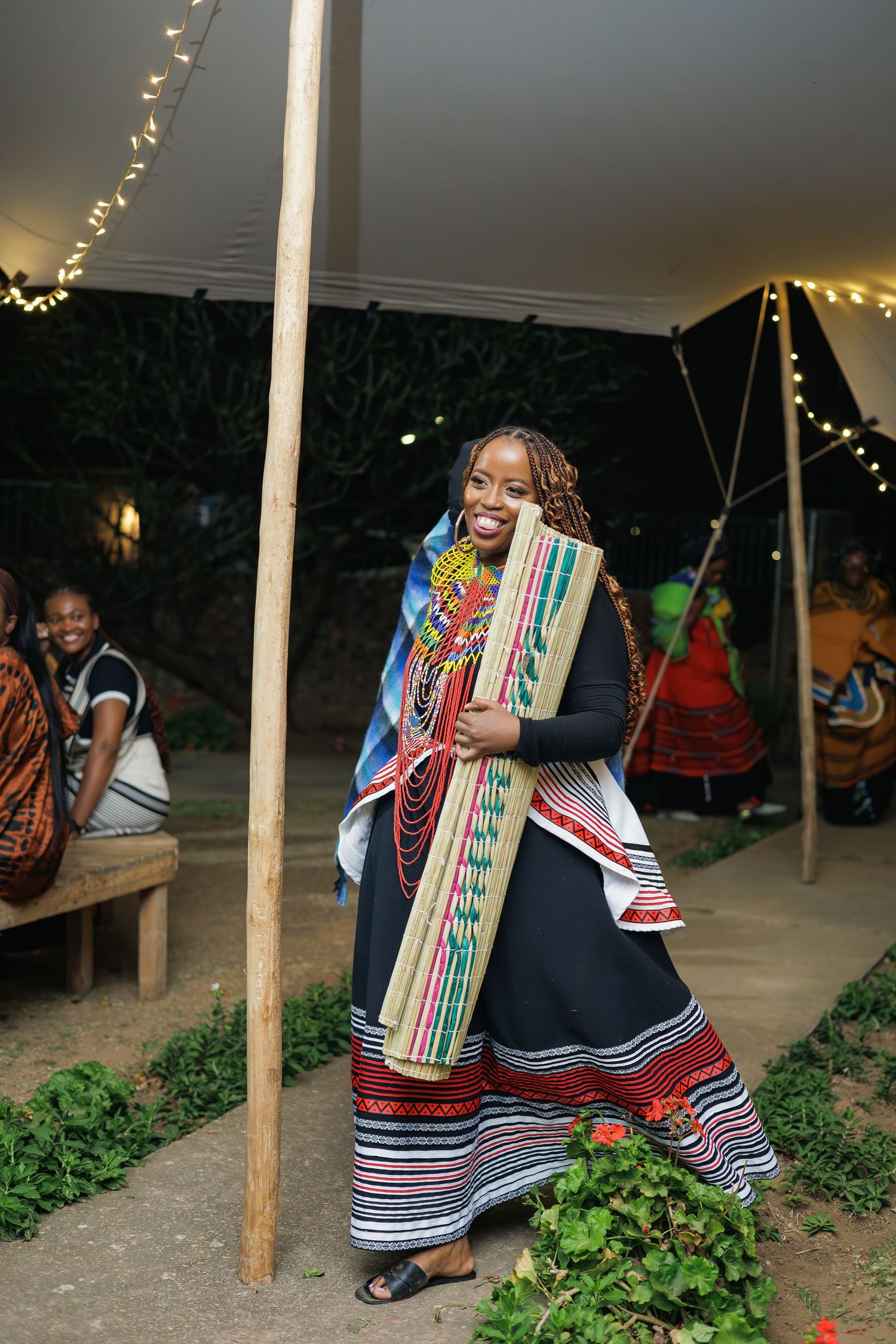 Smiling woman in traditional Xhosa dress carrying a woven mat at a nighttime cultural event.
