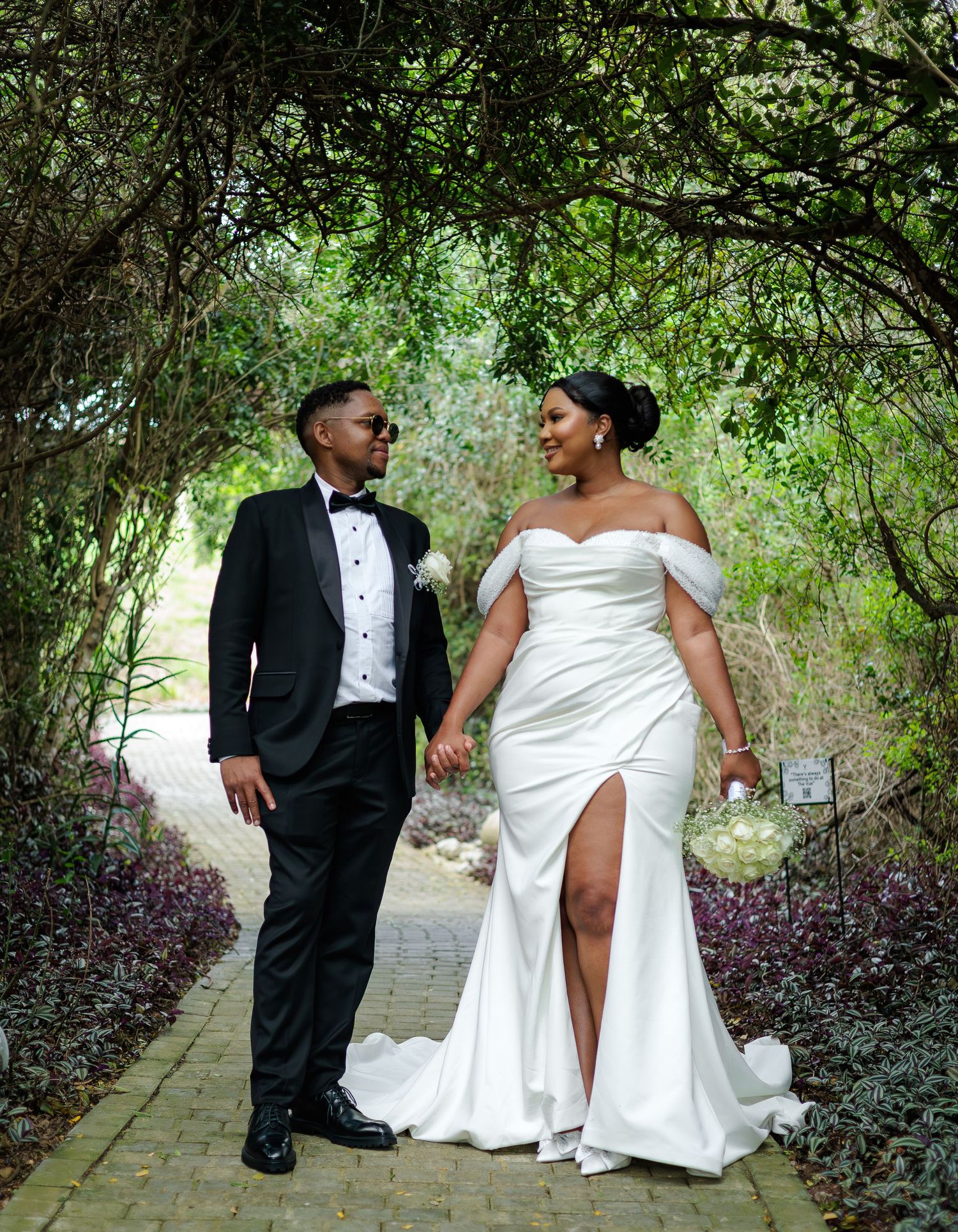 Bride and groom walking hand in hand under a lush green garden archway on their wedding day.
