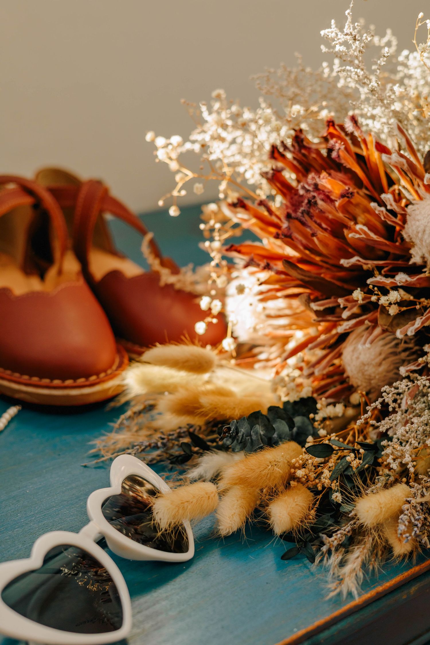 Boho wedding flat lay with dried flower bouquet, brown leather shoes, and heart-shaped sunglasses.