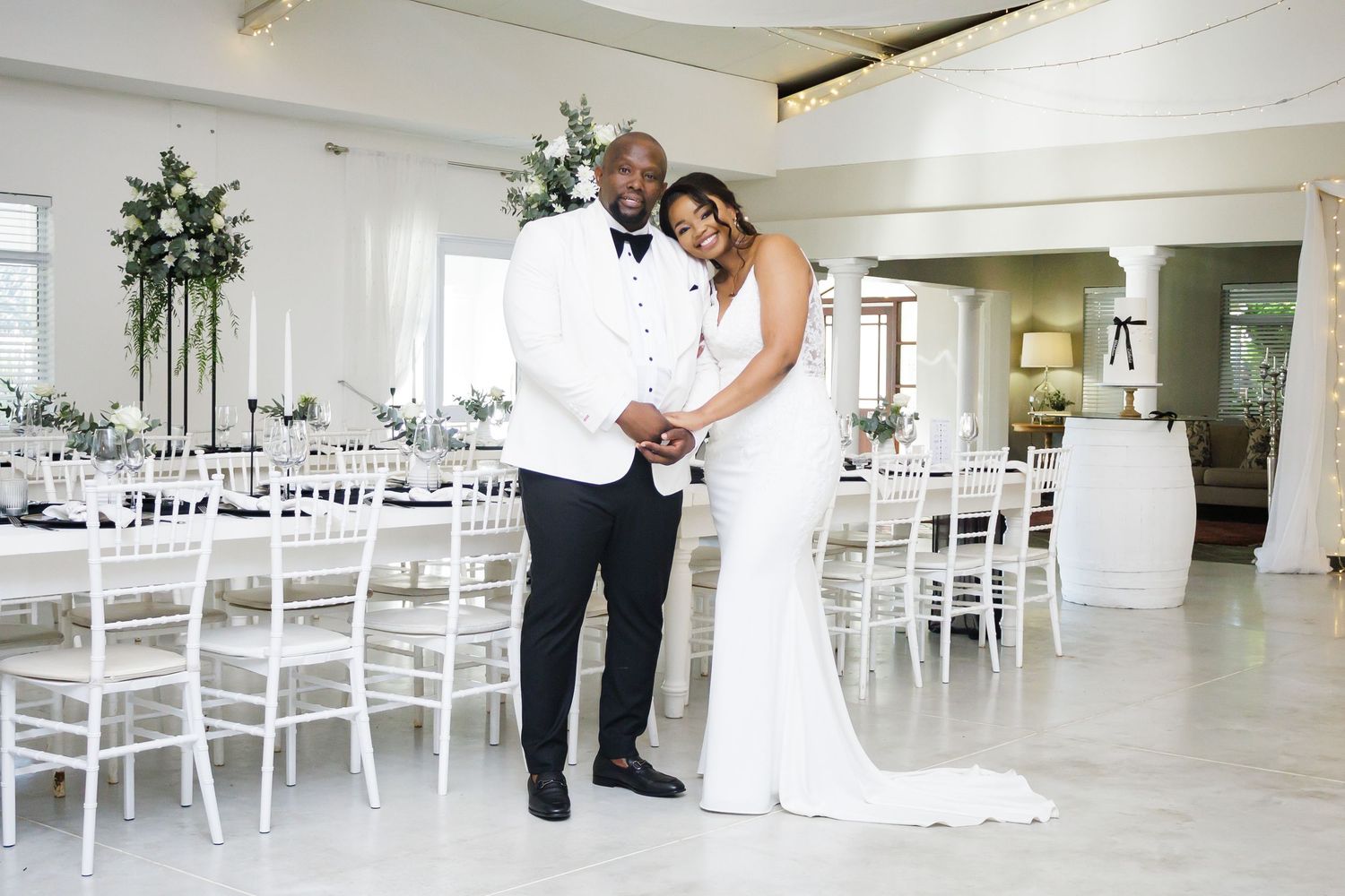 Elegant bride and groom posing in a white wedding reception venue with floral centerpieces.