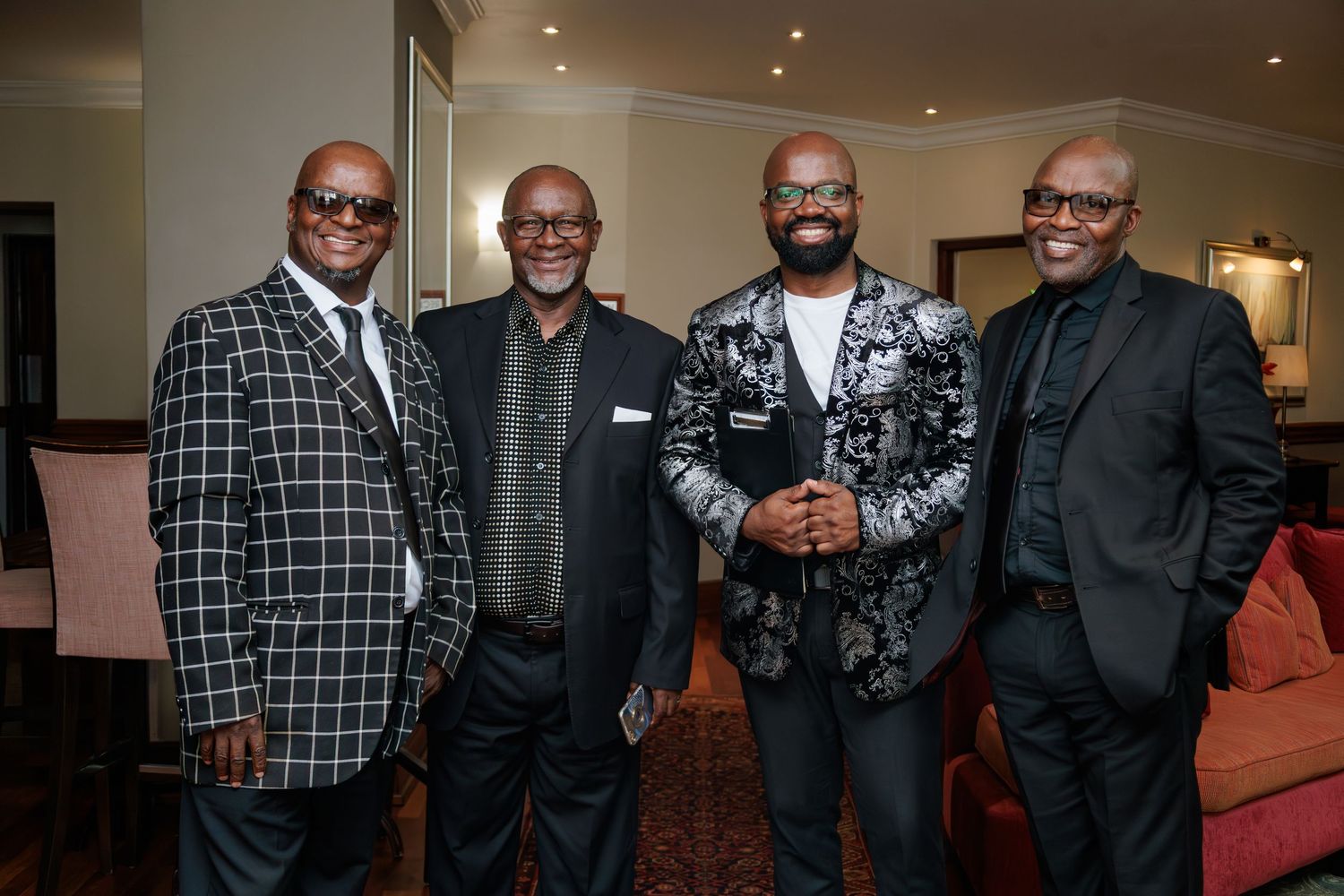 Four men smiling together in formal suits at an indoor event venue.