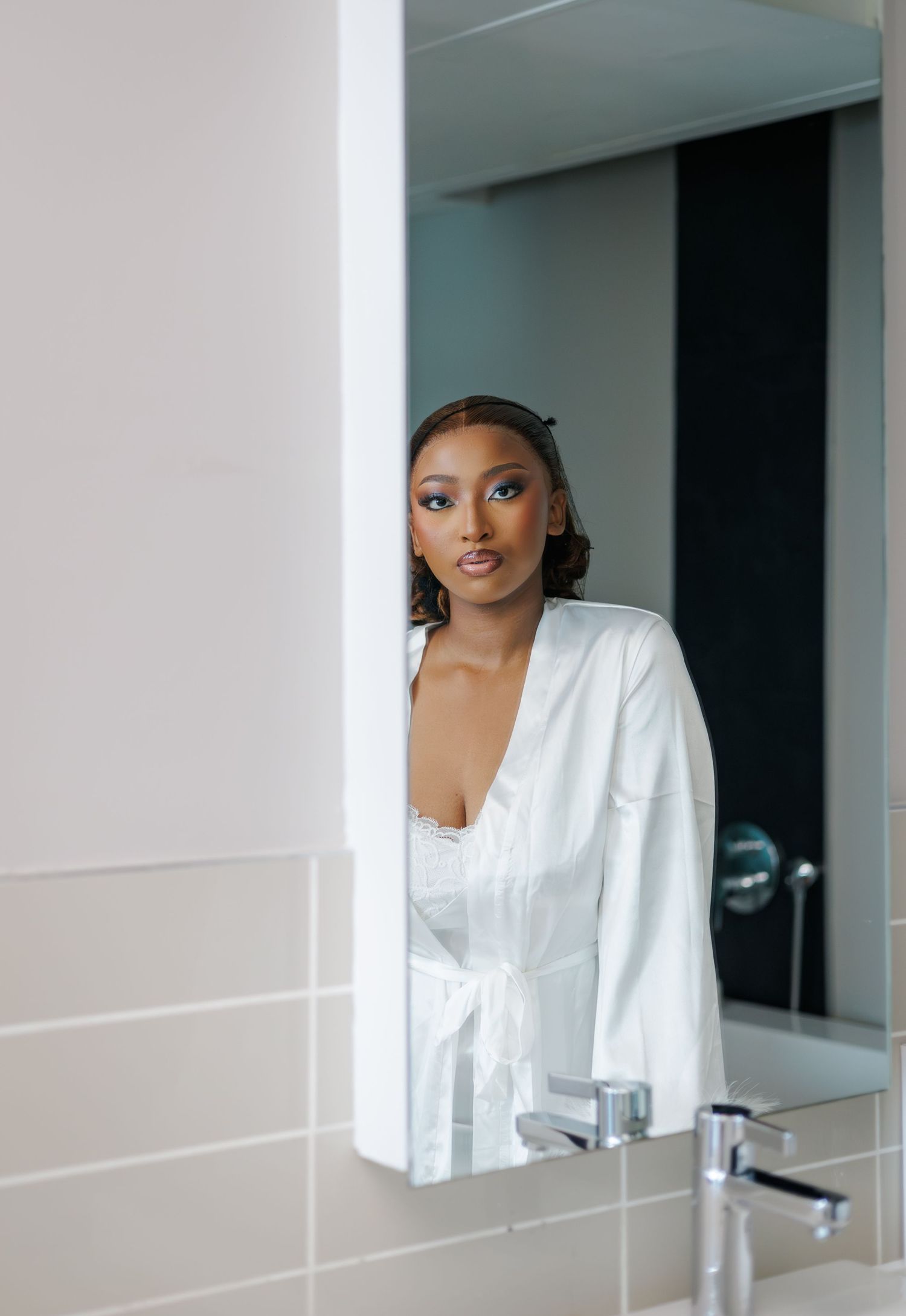 Elegant woman in white robe reflected in bathroom mirror with sophisticated makeup and updo.