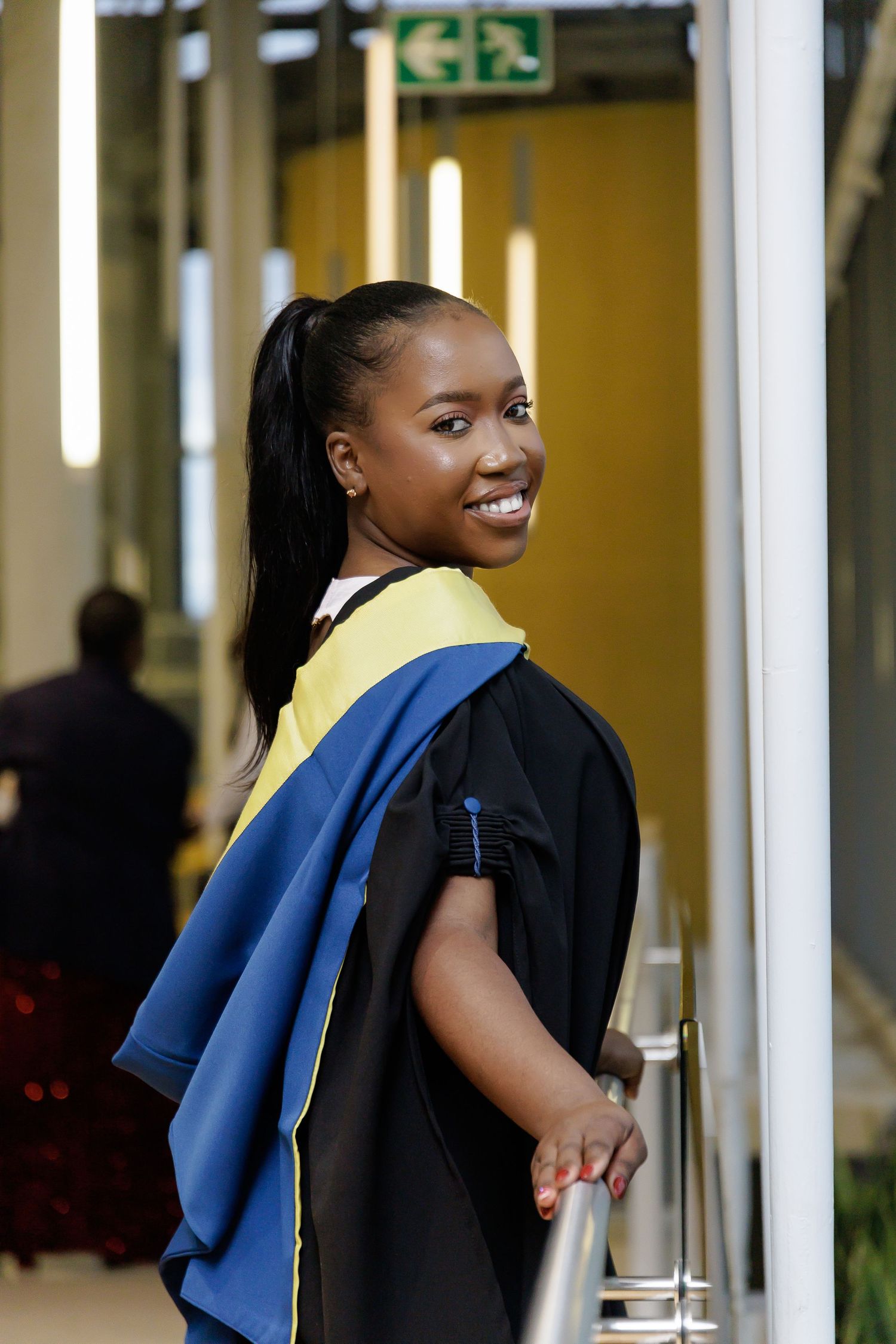 A smiling Black female graduate in black gown with yellow and blue hood poses by an indoor railing.