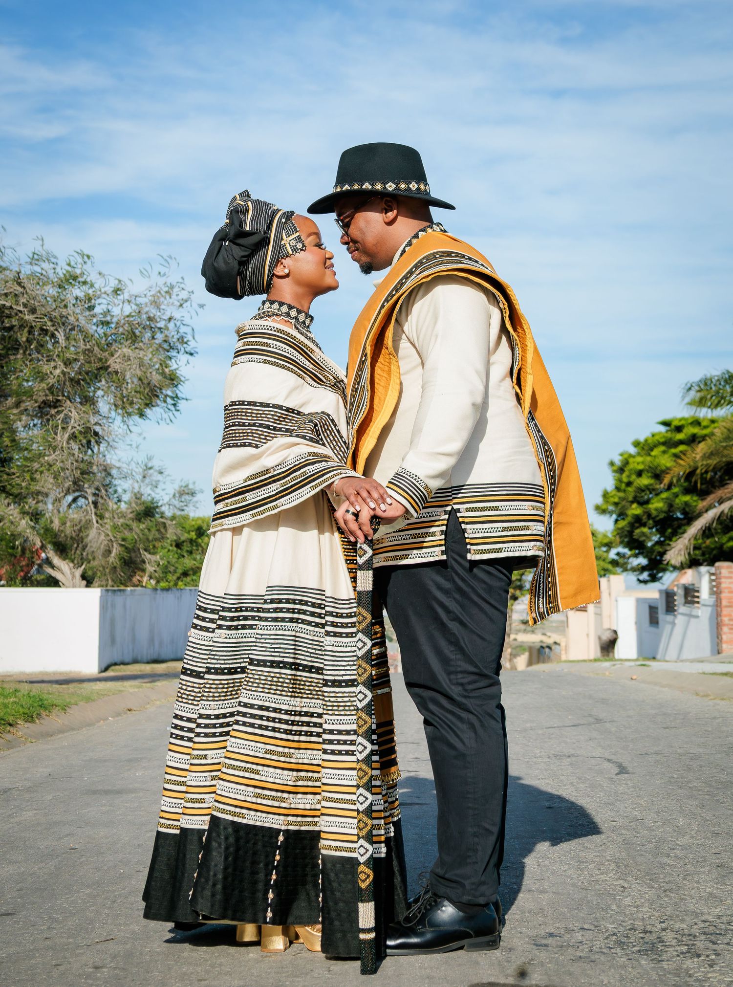 Couple in traditional Xhosa black and gold attire holding hands on a sunny street.