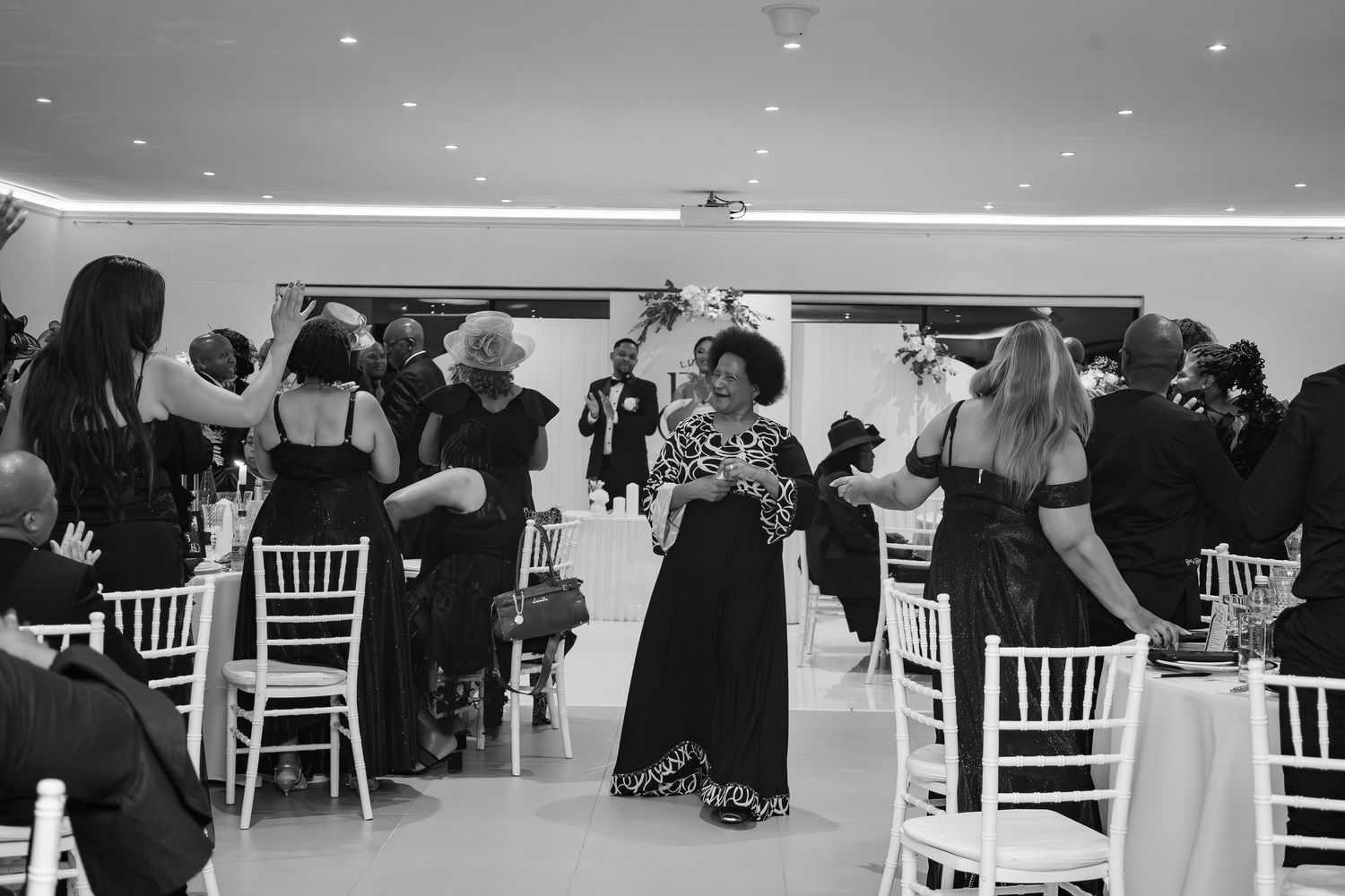 Black and white photo of a woman walking down the wedding ceremony aisle among seated guests.