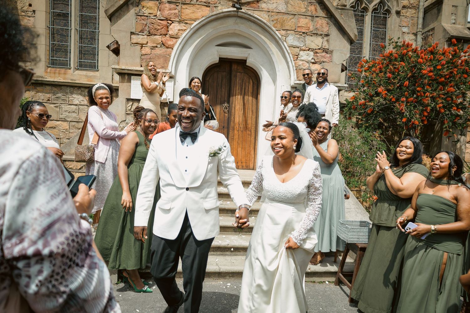 Newlyweds smile while exiting stone church surrounded by bridesmaids in sage green dresses.