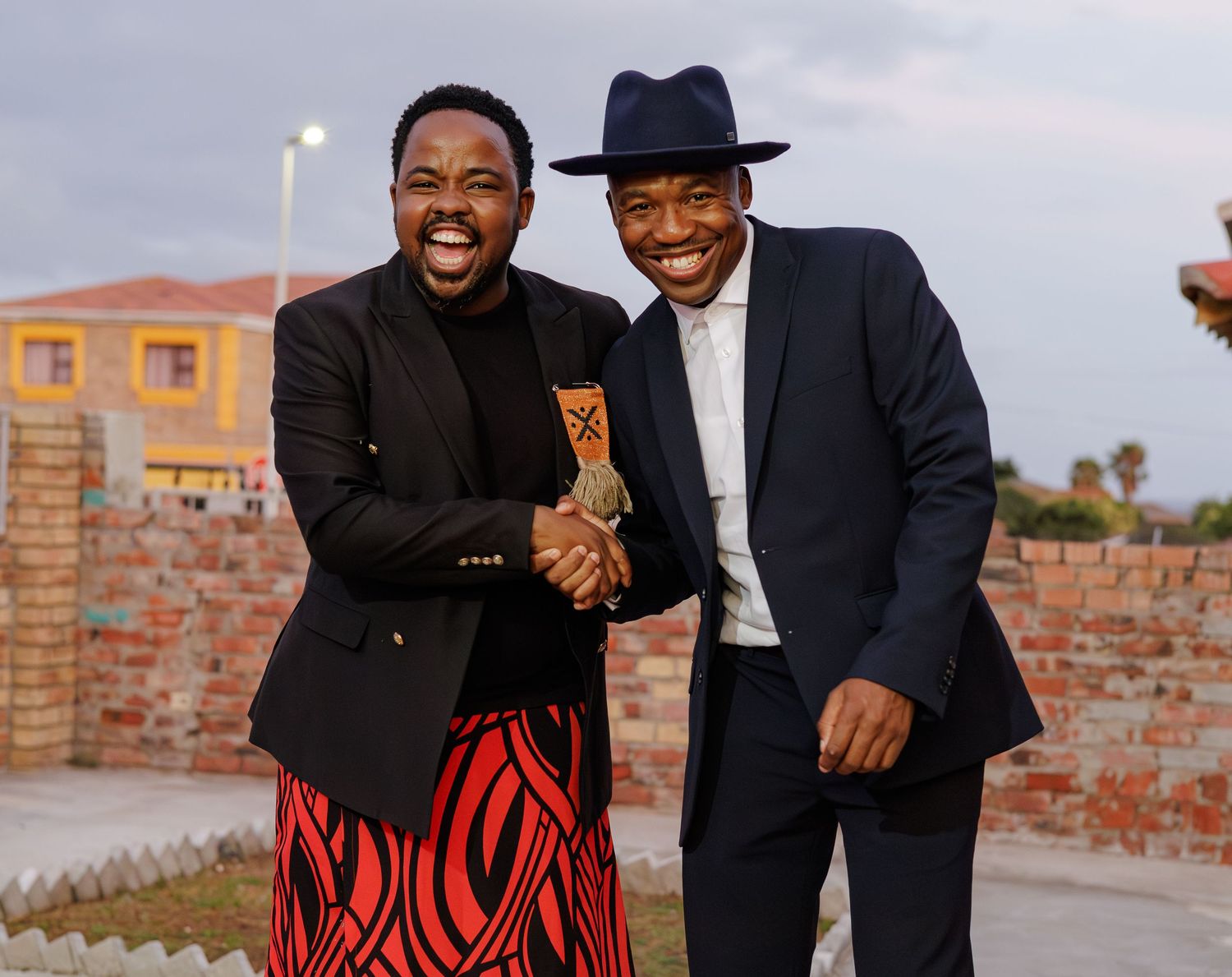 Two men in stylish attire shaking hands and smiling outdoors, one in black jacket with red skirt, other in navy suit.