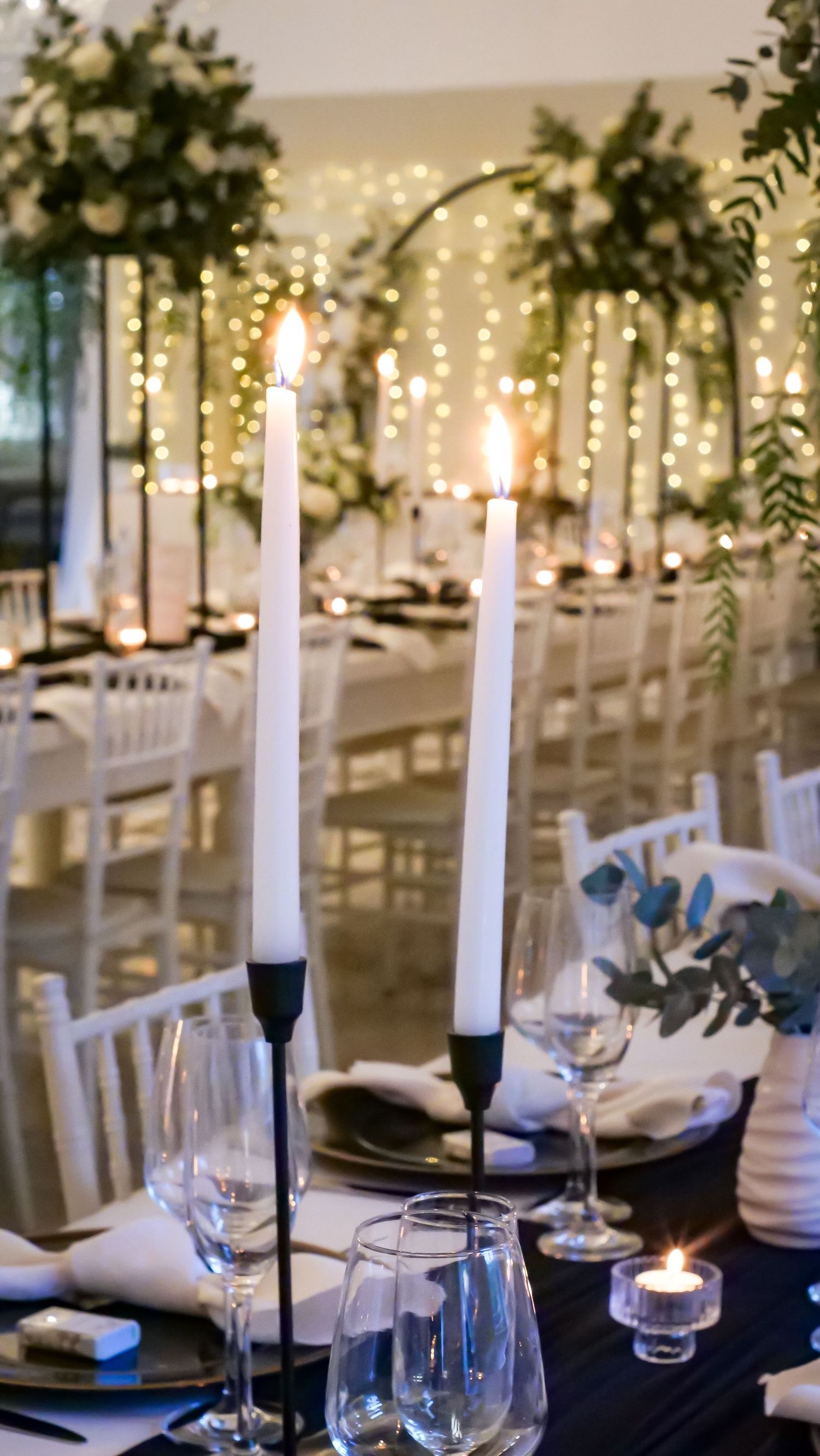 Elegant wedding table setting with white taper candles, glassware, and fairy light backdrop.