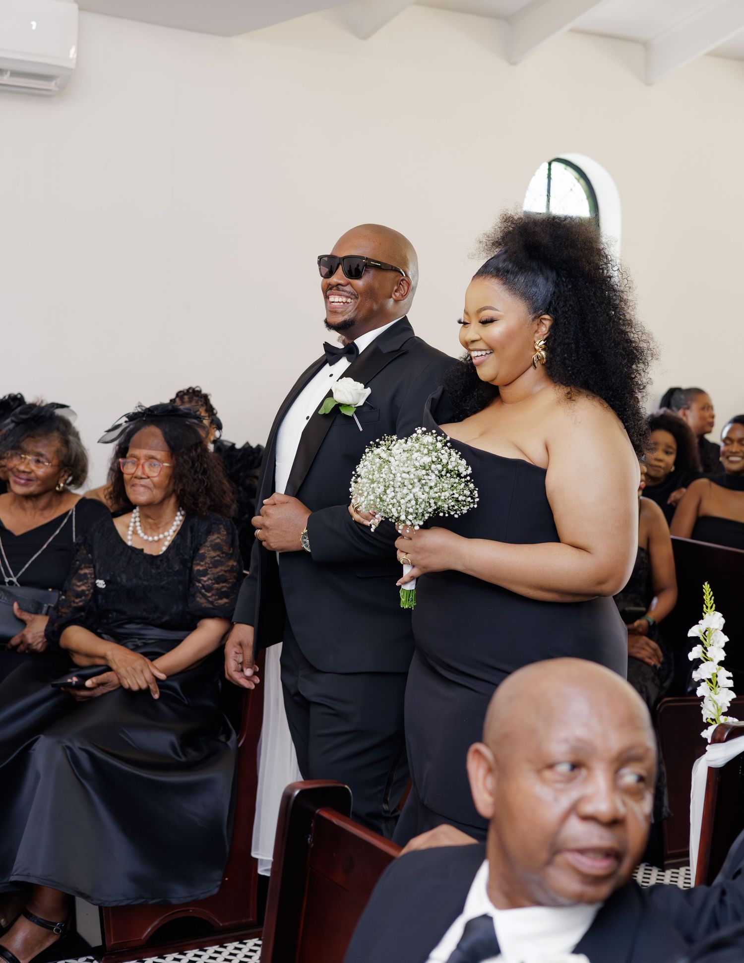 Groomsman in sunglasses and tuxedo escorts bridesmaid with baby's breath bouquet down aisle.