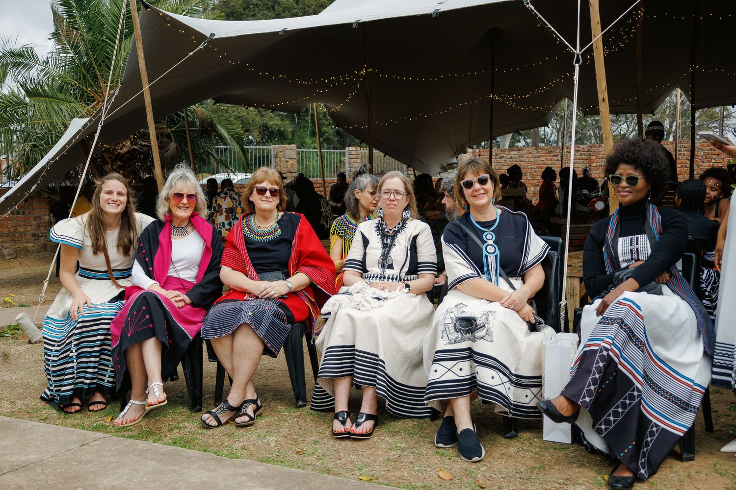Group of seven women wearing Xhosa-inspired clothing seated together under a tent at cultural event.