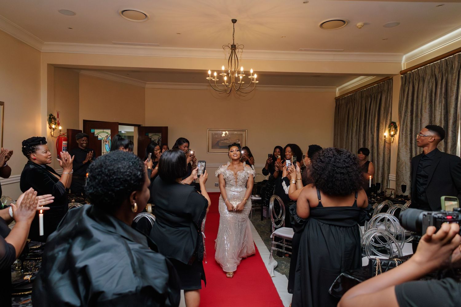 Bride in white gown walks red carpet entrance as guests applaud at elegant indoor wedding reception.