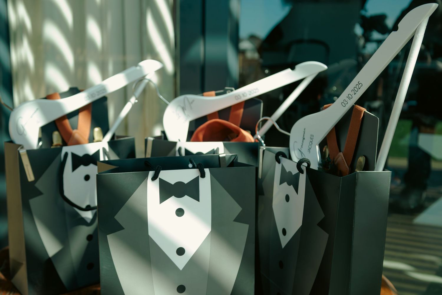 Wedding groomsmen gift bags with tuxedo design containing white hangers and accessories in sunlight.