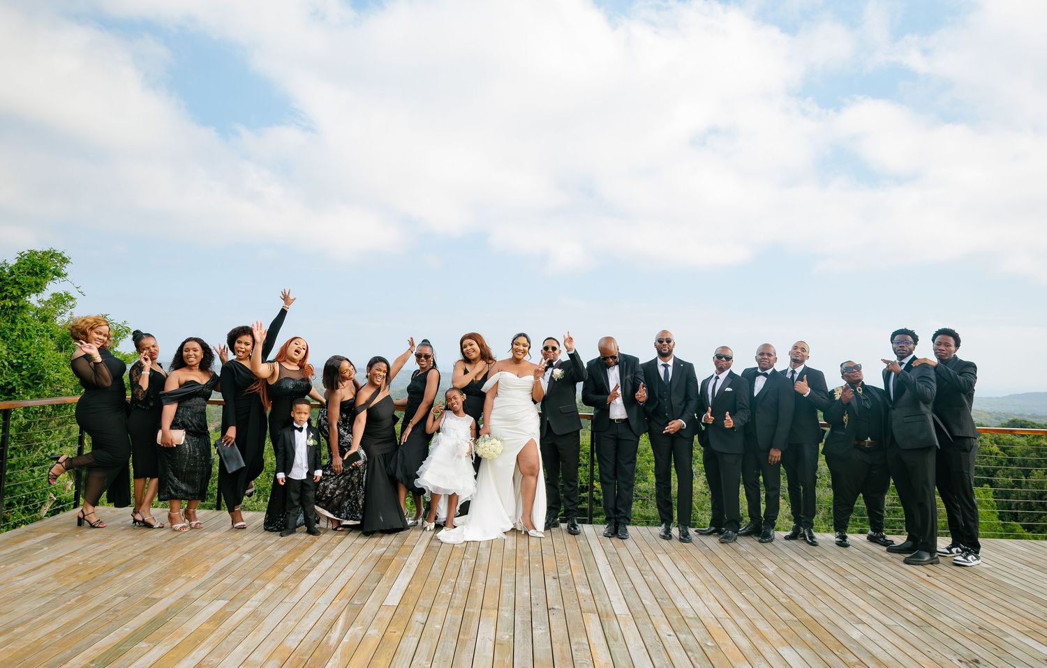 Large wedding party in black attire celebrating on a wooden deck with scenic sky views.