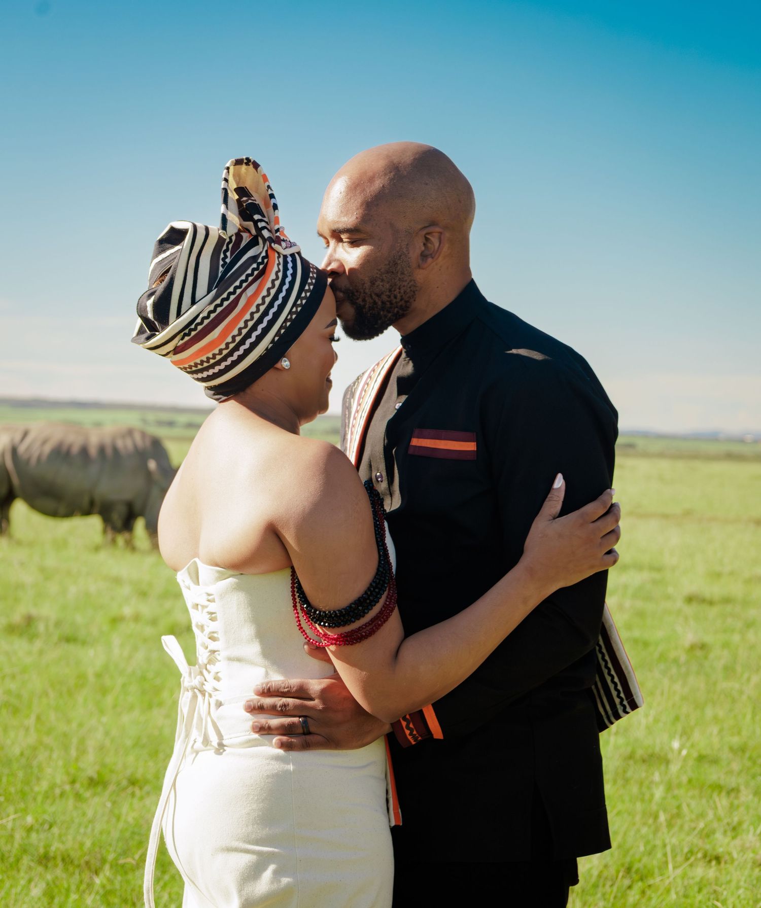 Couple in traditional African attire sharing a kiss in a green field, with a rhino visible in the background.